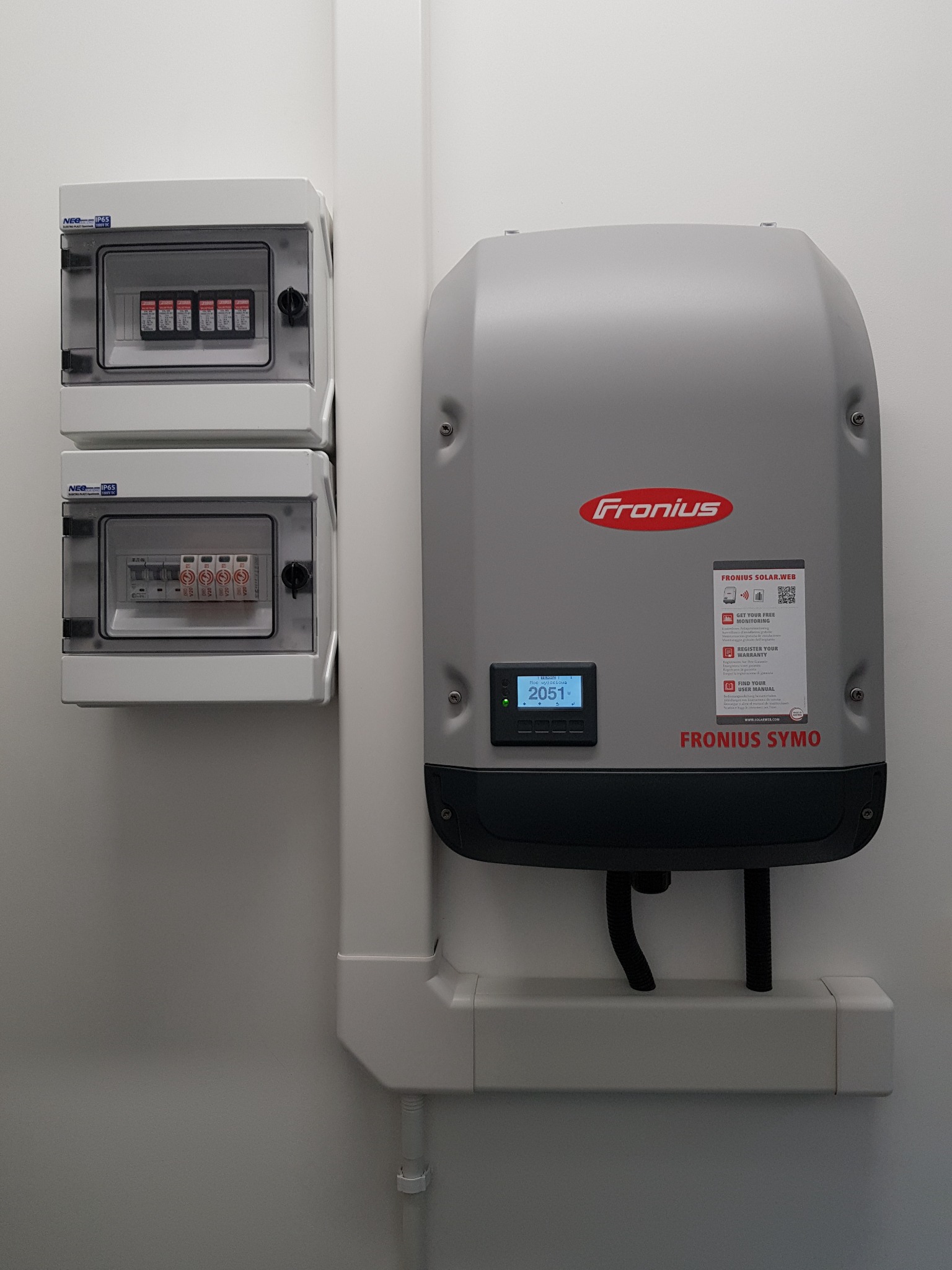Szare skrzynki bezpiecznikowe oraz falownik Fronius Symo z wyświetlaczem wskazującym moc 2051W zamontowane na białej ścianie, widoczne przewody i plastikowe korytko kablowe.