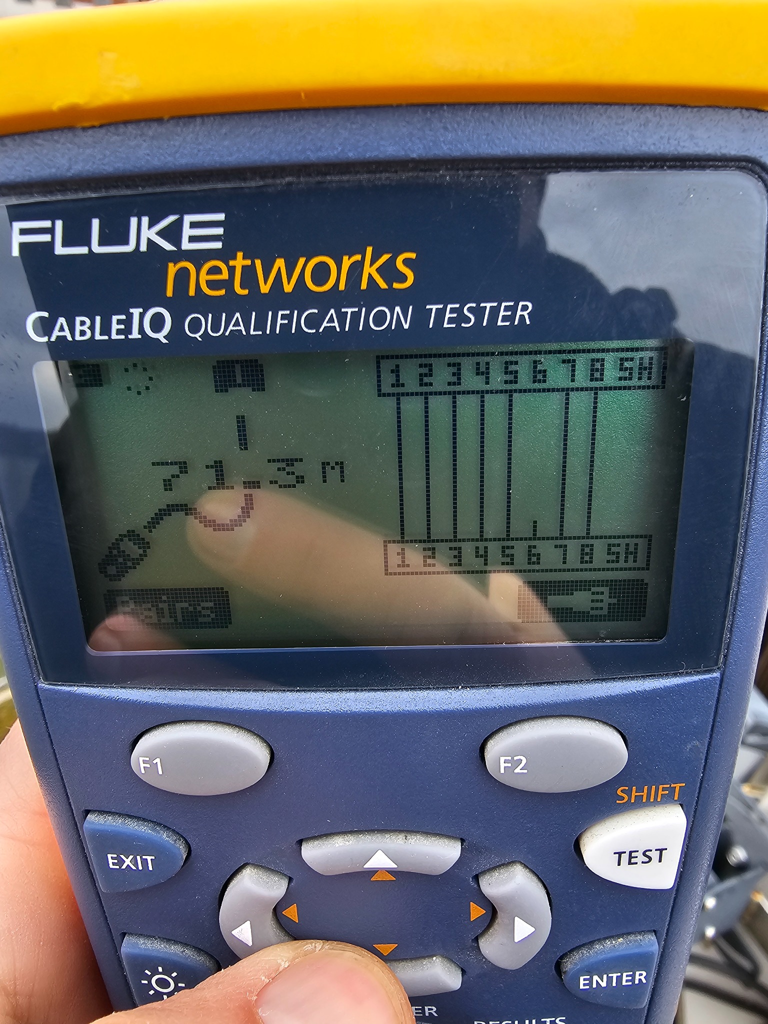 Tester okablowania Fluke Networks CableIQ Qualification Tester z widocznym ekranem LCD wskazującym wynik pomiaru 71.3m oraz diagramem par kablowych, w tle odbicie dłoni operatora.