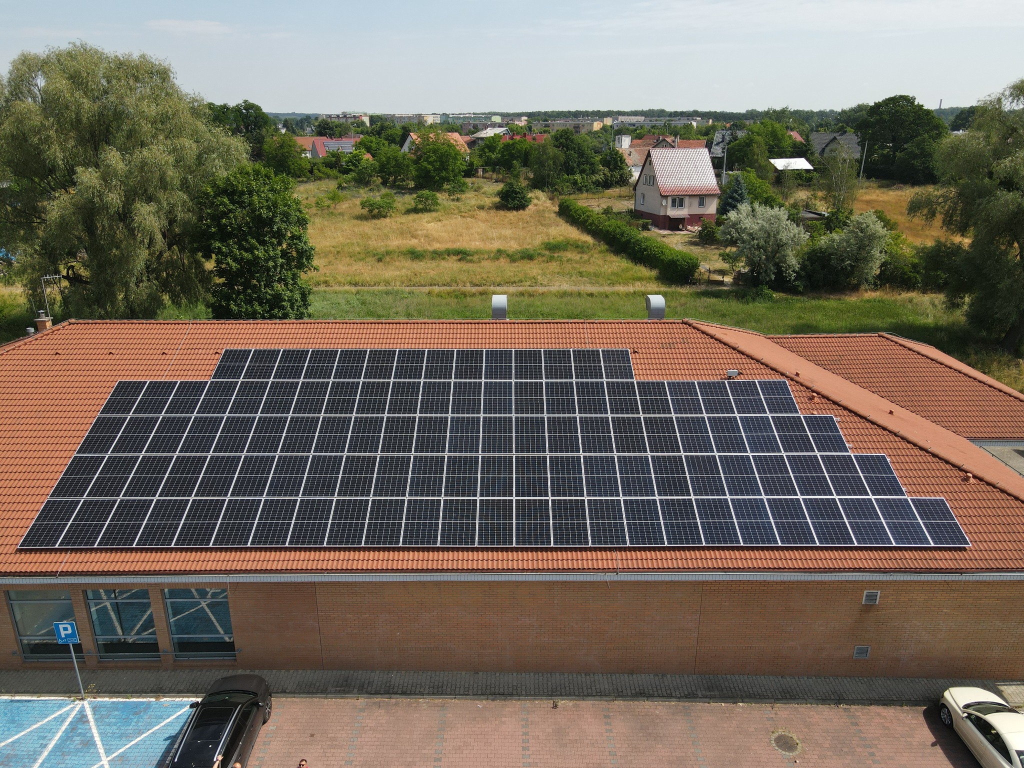 Widok z góry na budynek z panelami fotowoltaicznymi na dachu w Wołowie, z widocznym parkingiem i otaczającą zielenią.