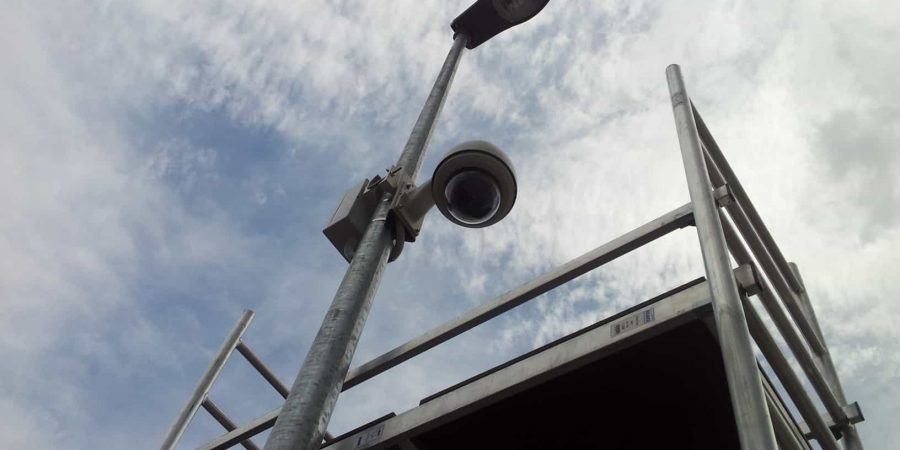 Kamera kopułkowa zamontowana na słupie oświetleniowym, widoczna z dołu, z metalową konstrukcją rusztowania w tle i pochmurnym niebem.