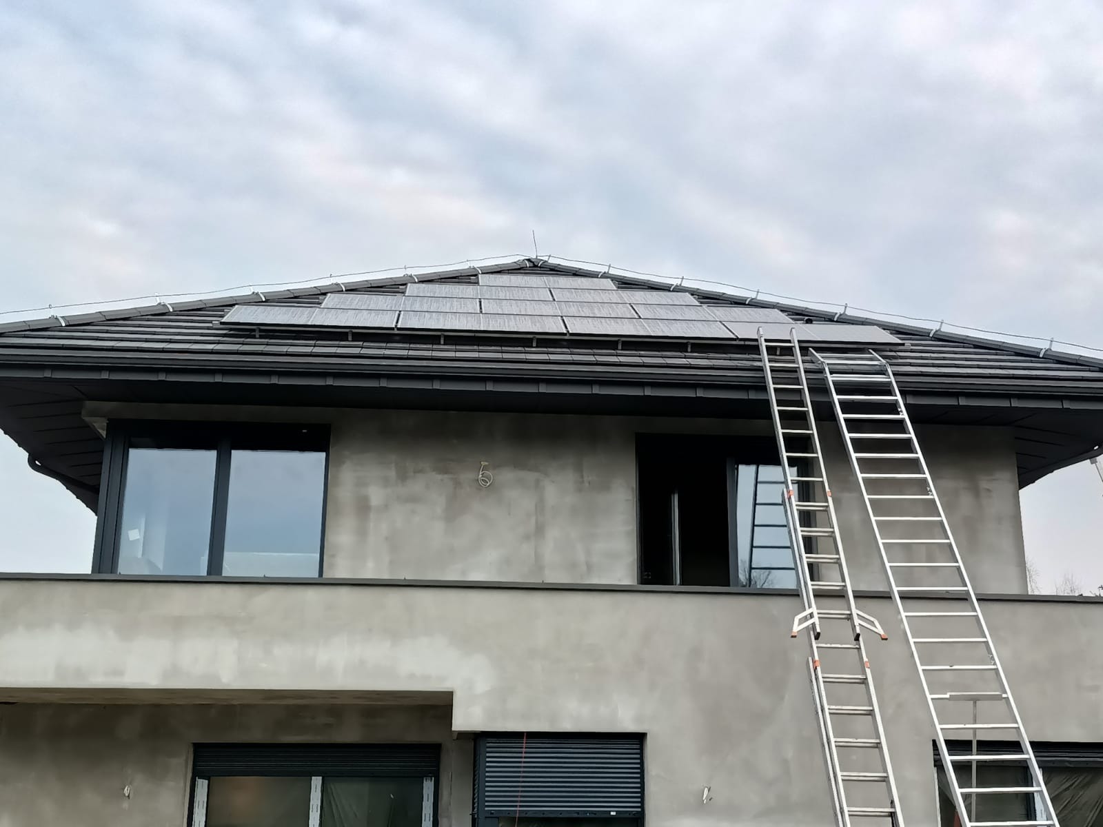 Szereg paneli słonecznych na dachu domu jednorodzinnego w trakcie montażu, widoczne dwie aluminiowe drabiny opierające się o dach, elewacja budynku w stanie surowym.