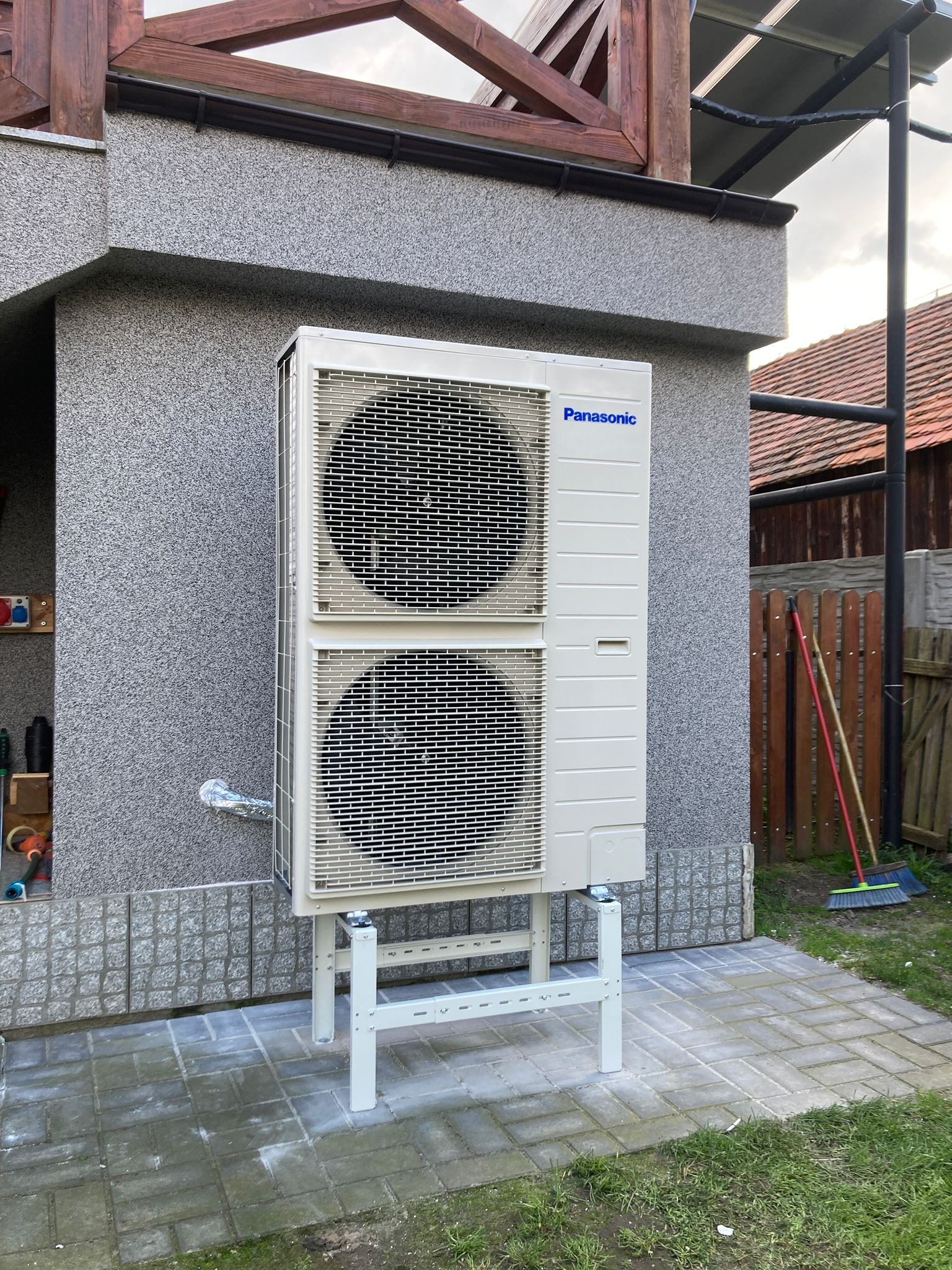 Biała, dwusekcyjna pompa ciepła marki Panasonic zamontowana na metalowym stelażu przy ścianie budynku z szarą elewacją i drewnianą konstrukcją dachu.
