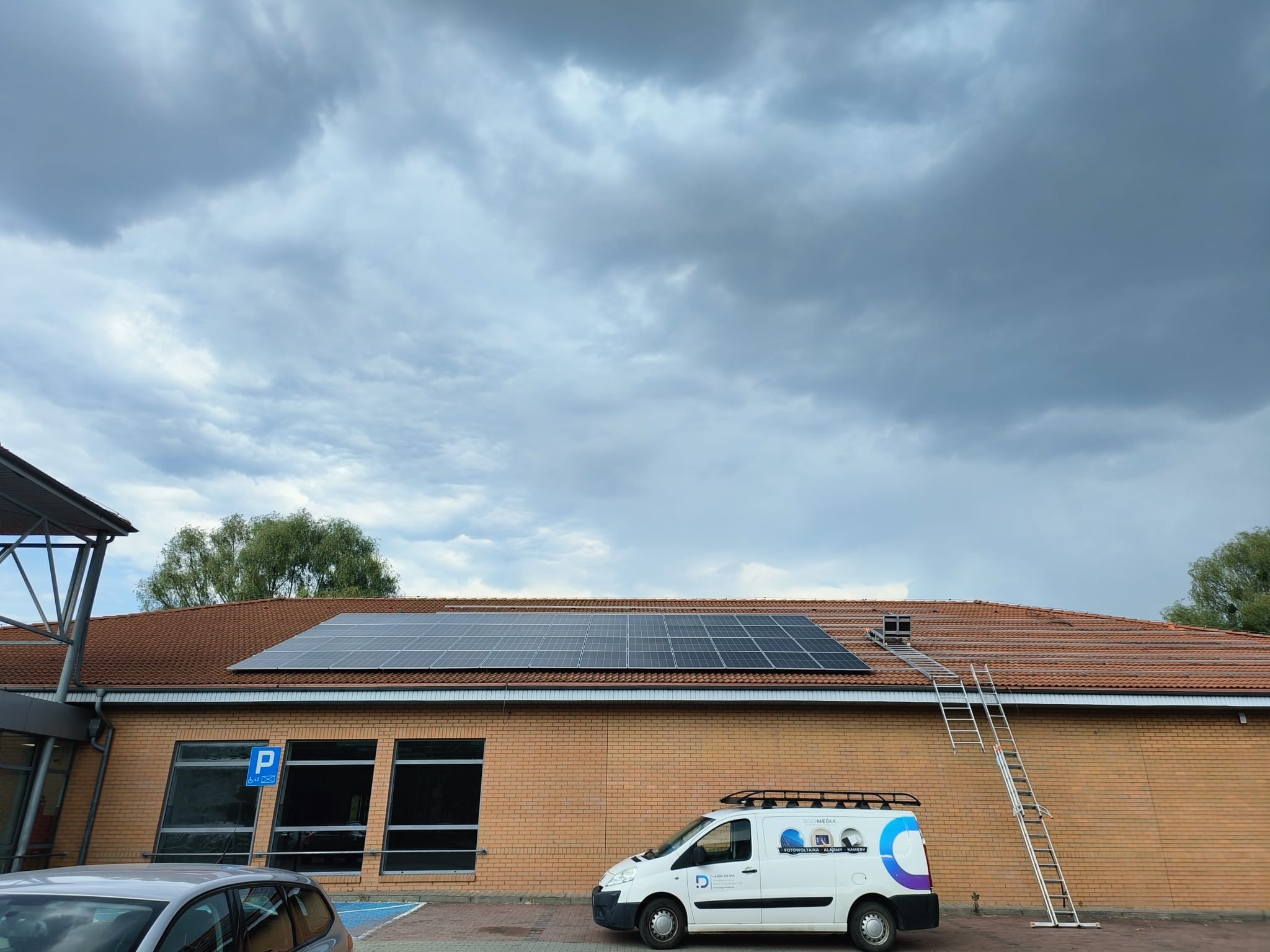 Panel słoneczny zainstalowany na dachu budynku z cegły, obok zaparkowany biały samochód dostawczy z logo firmy, drabina oparta o dach, pochmurne niebo w tle.