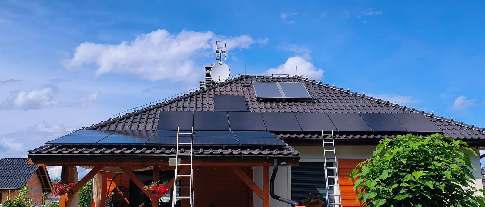 Czarny panel fotowoltaiczny na brązowym dachu domu jednorodzinnego z drabinkami opartymi o dach, błękitne niebo z białymi chmurami w tle.