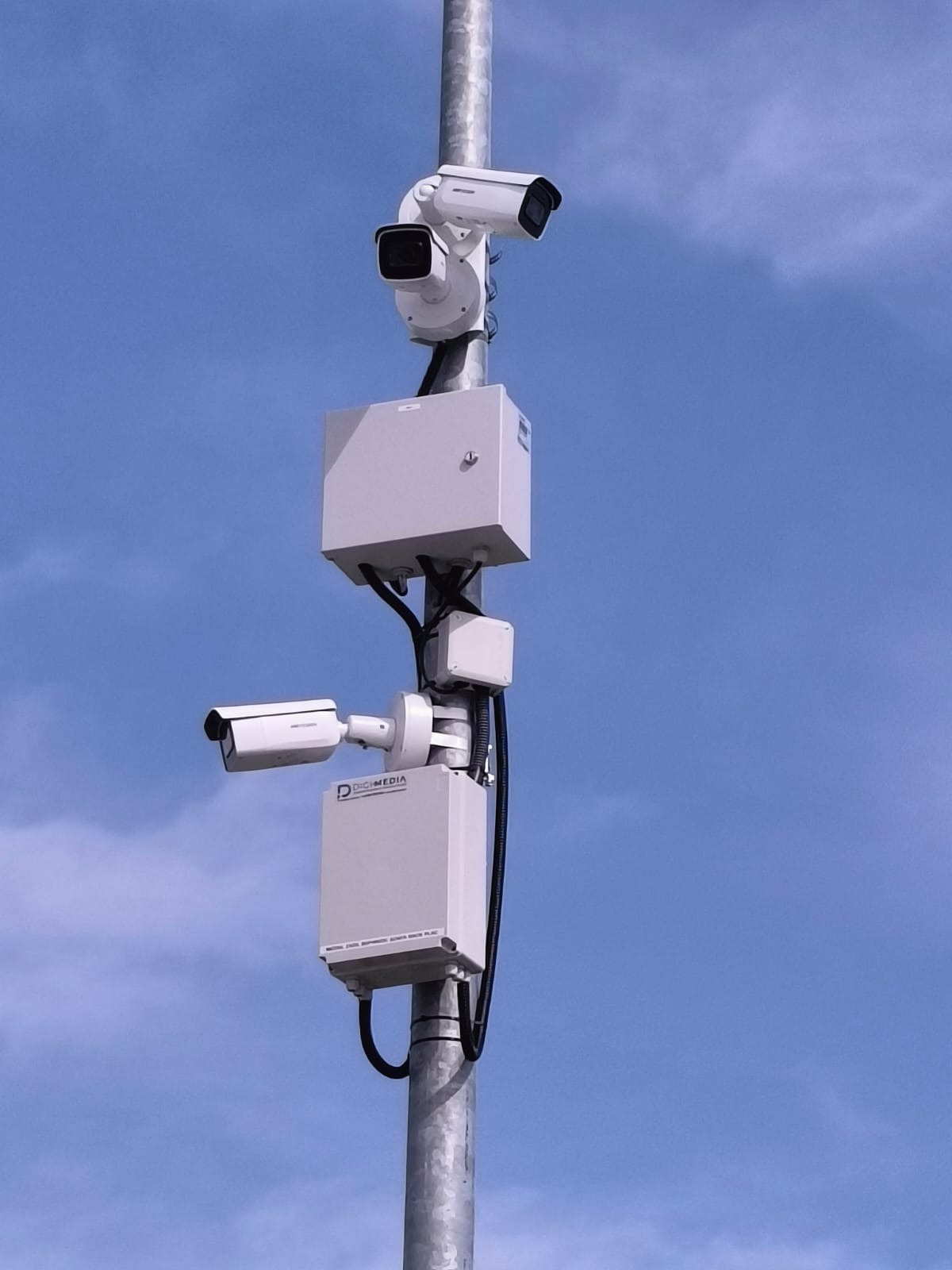 Dwie kamery monitoringu na metalowym słupie, jedna nad drugą, z dodatkowymi skrzynkami sterującymi i okablowaniem na tle błękitnego nieba.