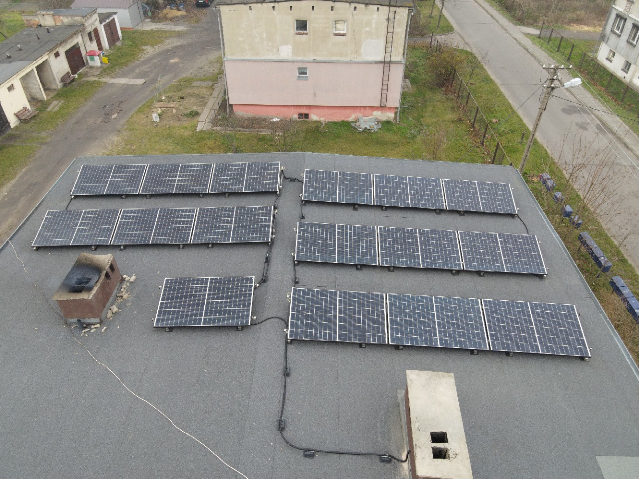 Widok z góry na instalację paneli słonecznych na dachu budynku z dwoma kominami. Panele ułożone w rzędach, widoczne okablowanie. W tle budynki mieszkalne i droga.