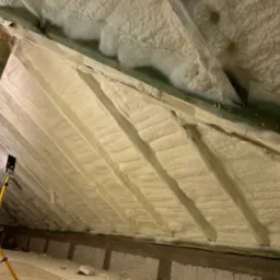 Poddasze ocieplone pianką PUR, widoczne drewniane elementy konstrukcyjne i lampa na statywie. Aplikacja pianki zakończona, gotowa izolacja termiczna.