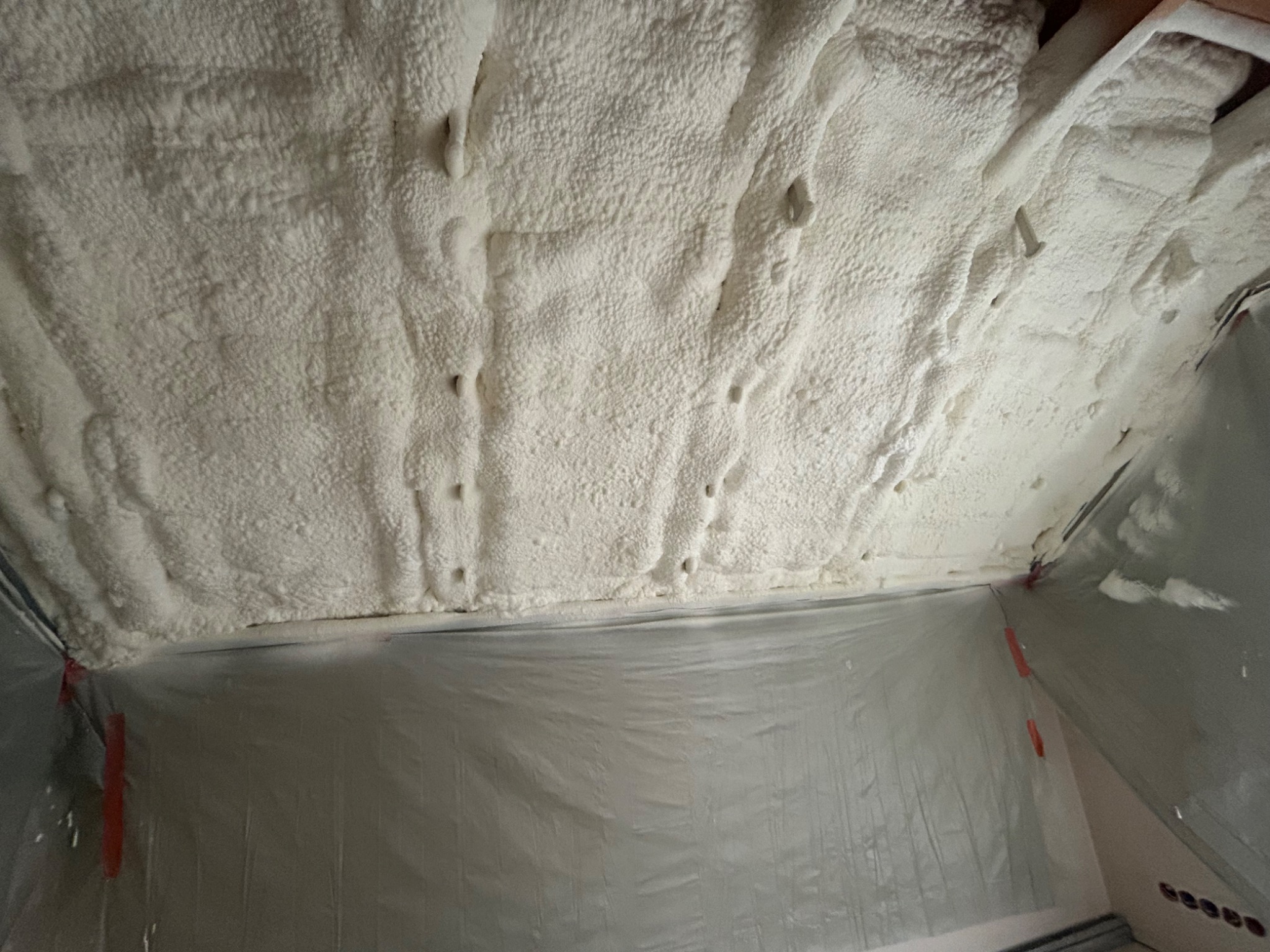 Ocieplenie poddasza pianką PUR: widok z dołu na białą, nierówną strukturę pianki na suficie, zabezpieczoną folią. Widoczne punkty mocowania pianki.