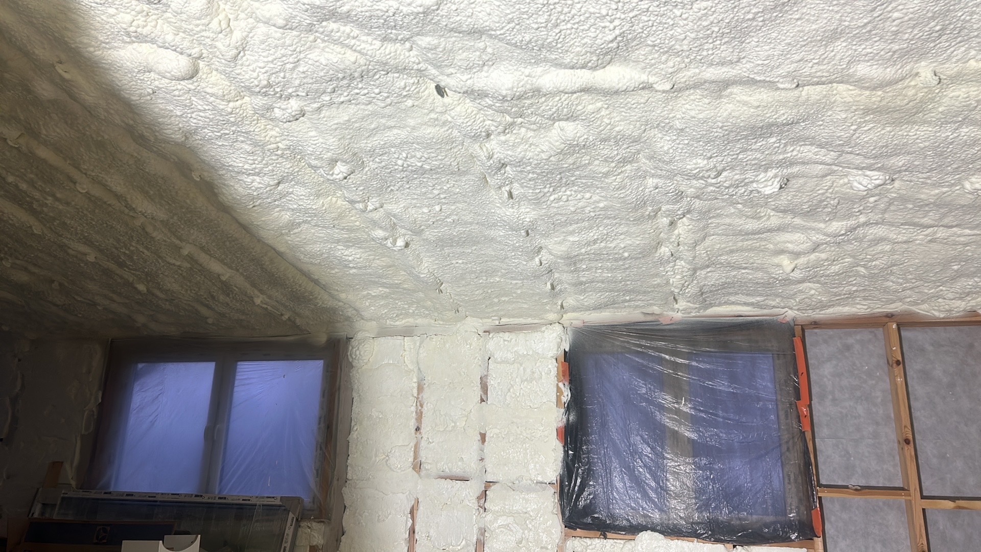 Ocieplenie poddasza pianką PUR: widok na sufit i ściany pokryte grubą warstwą białej piany, okna zabezpieczone folią. Detal: nierównomierna struktura pianki.