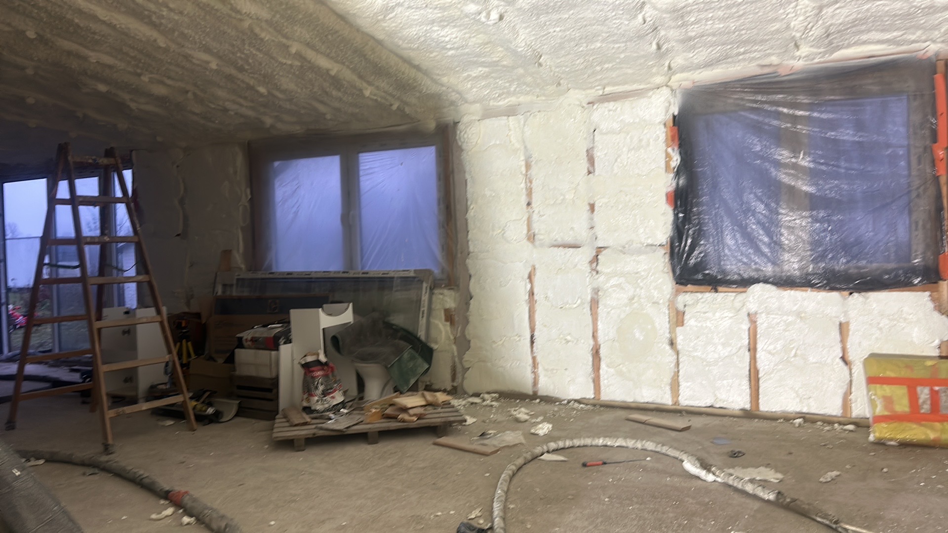 Wnętrze w trakcie ocieplania: ściany i sufit pokryte pianką PUR, okna zabezpieczone folią, drabina oparta o ścianę. Widoczne elementy instalacji.