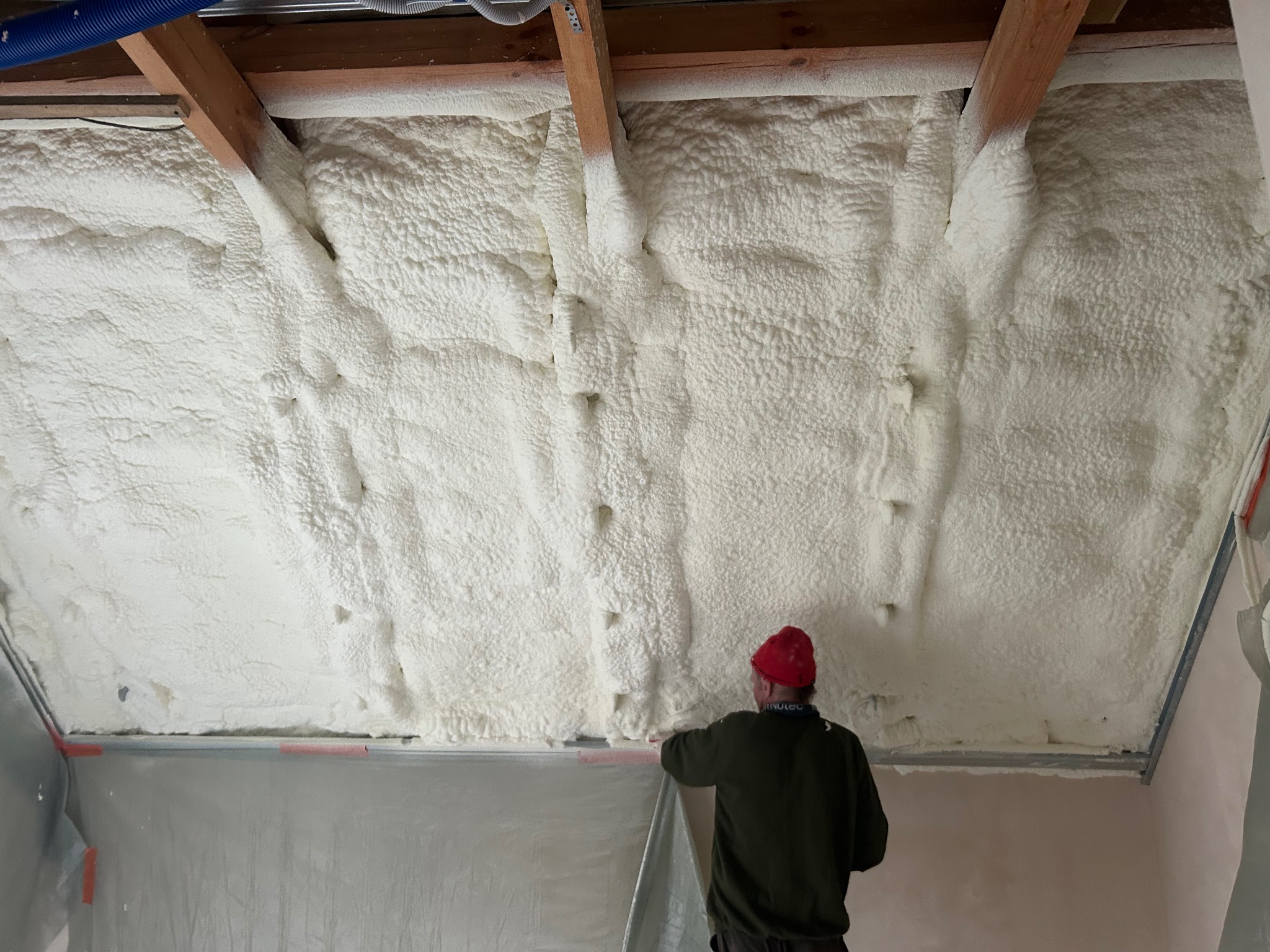 Poddasze z drewnianą konstrukcją pokryte białą pianką PUR, widoczny pracownik w czerwonej czapce podczas aplikacji. Izolacja natryskowa.