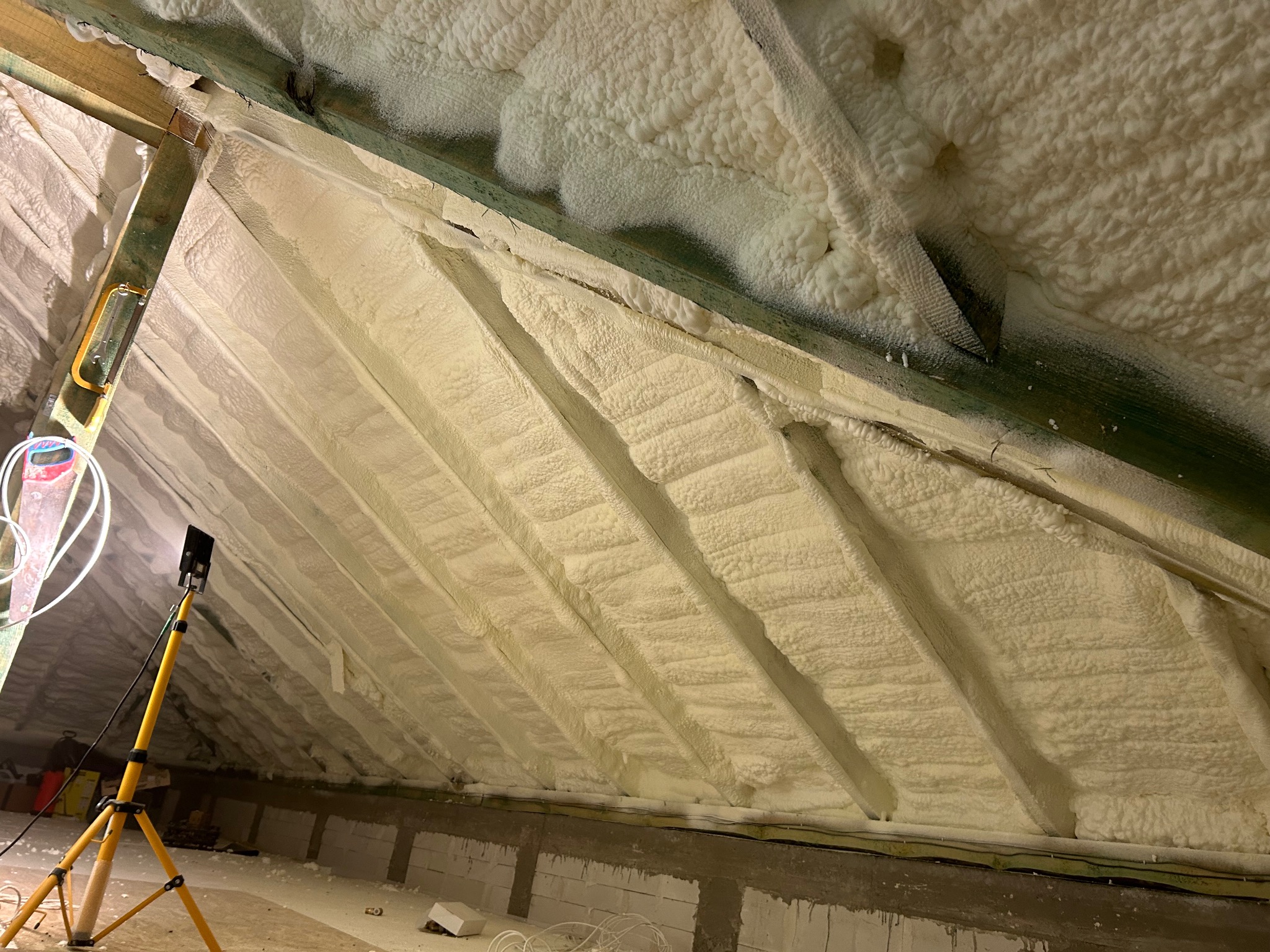 Poddasze ocieplone pianką PUR, widoczne drewniane elementy konstrukcyjne i lampa na statywie. Aplikacja pianki zakończona, gotowa izolacja termiczna.