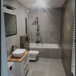 Kamil Talarek - Nowoczesna łazienka w szarościach z drewnianym blatem pod umywalką, czarna armatura, płytki imitujące kamień, pralka wkomponowana w zabudowę.