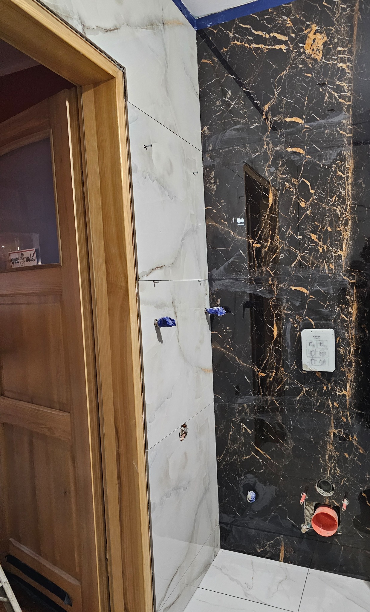 Ściana łazienki w trakcie układania płytek: białe płytki imitujące marmur sąsiadują z czarnymi płytkami z wyraźnym złotym żyłkowaniem, widoczne niebieskie kliny dystansowe, zamontowany stelaż...