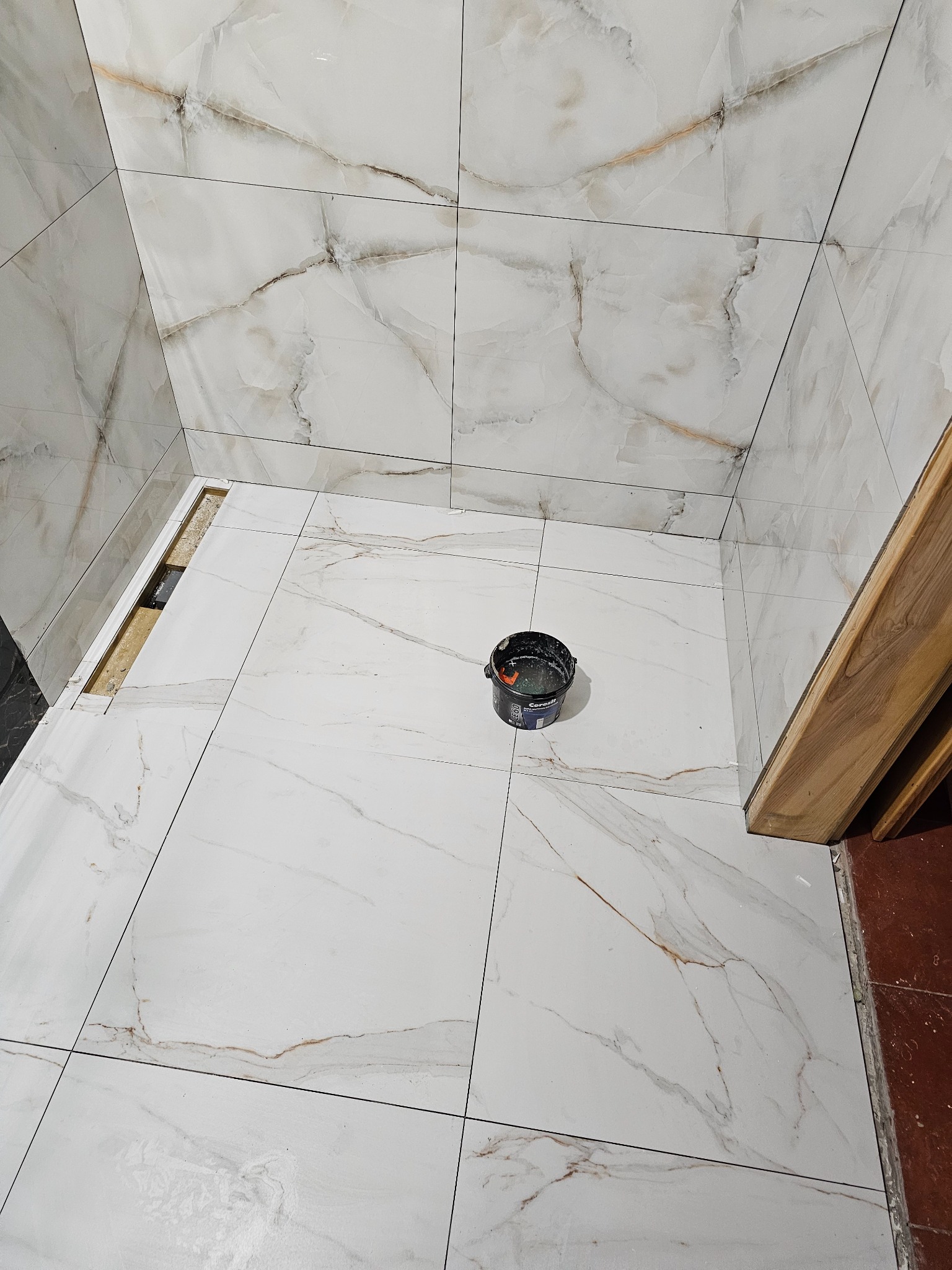 Układanie białych płytek imitujących marmur w łazience w Maszewie, widoczny odpływ liniowy i wiaderko z zaprawą.