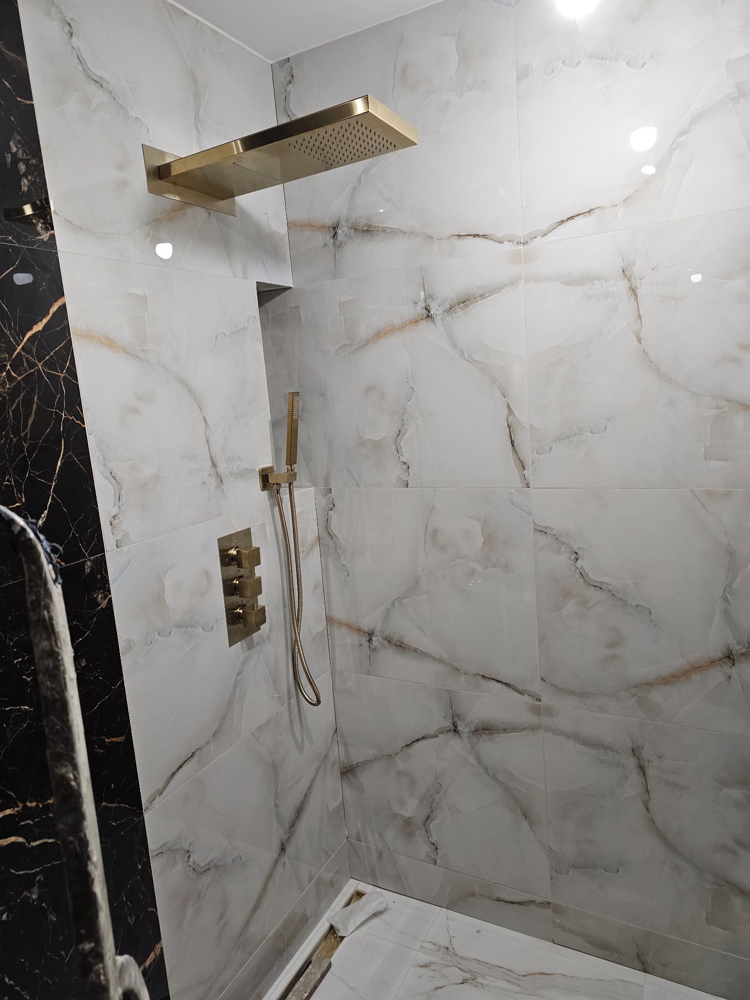 Nowoczesna kabina prysznicowa z białymi płytkami imitującymi marmur z brązowymi żyłami, armatura w kolorze szczotkowanego złota: deszczownica, bateria podtynkowa i słuchawka prysznicowa.