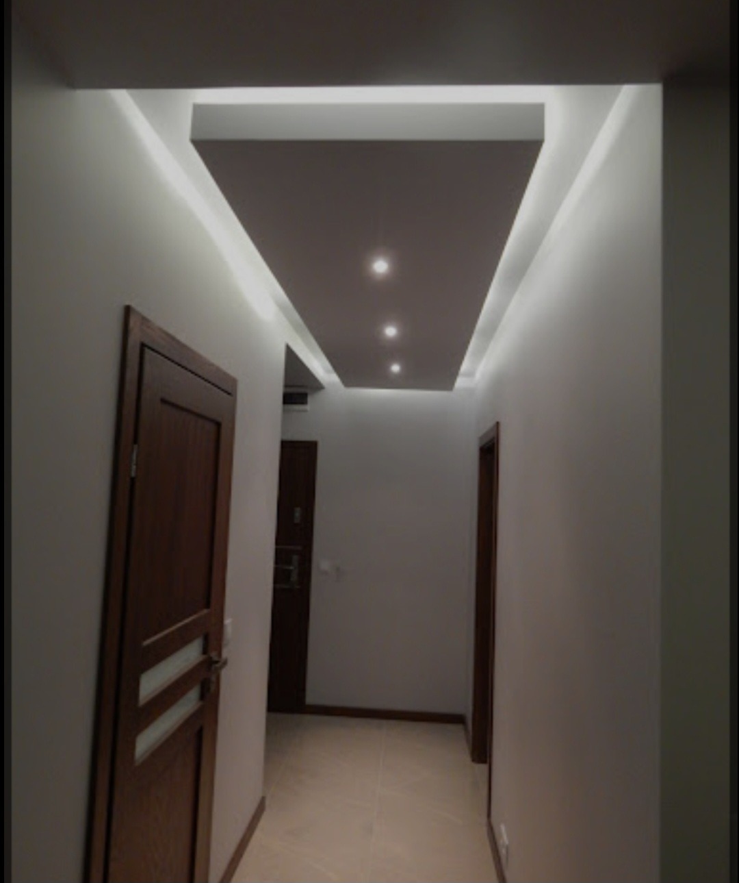 Korytarz z podwieszonym sufitem z oświetleniem punktowym i ukrytym oświetleniem LED, widoczne dwoje drzwi z ciemnego drewna.