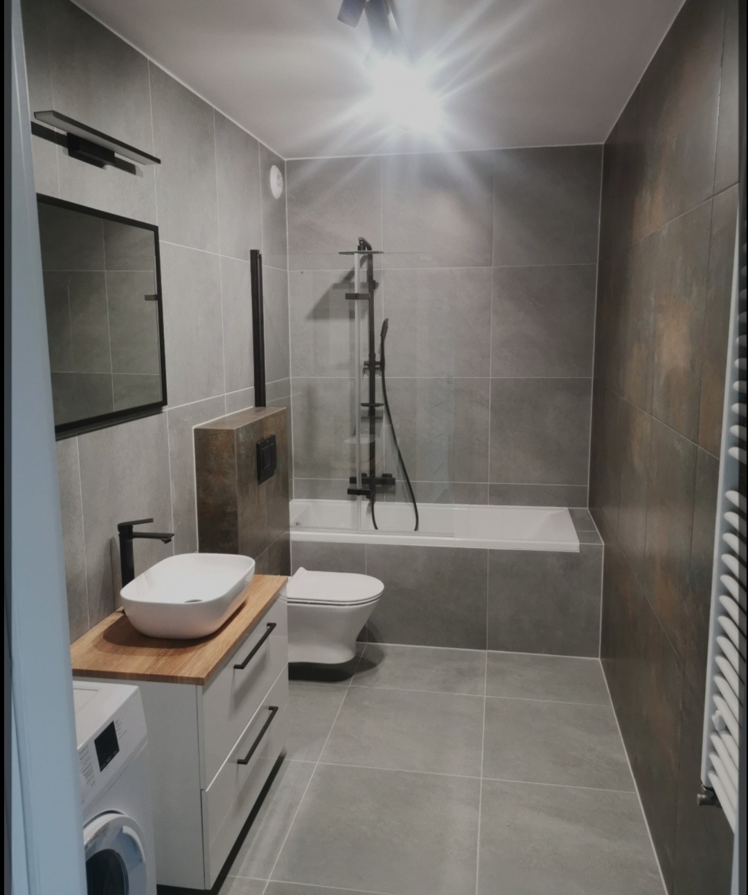 Nowoczesna łazienka w szarościach z drewnianym blatem pod umywalką, czarna armatura, płytki imitujące kamień, pralka wkomponowana w zabudowę.