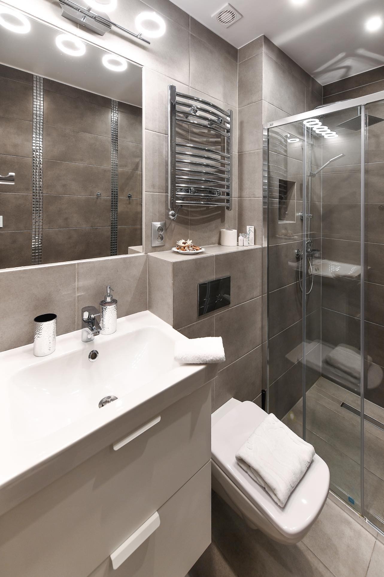 Nowoczesna łazienka w Warszawie z kabiną prysznicową, grzejnikiem drabinkowym i umywalką z szafką, wykończona szarymi płytkami o matowej powierzchni.