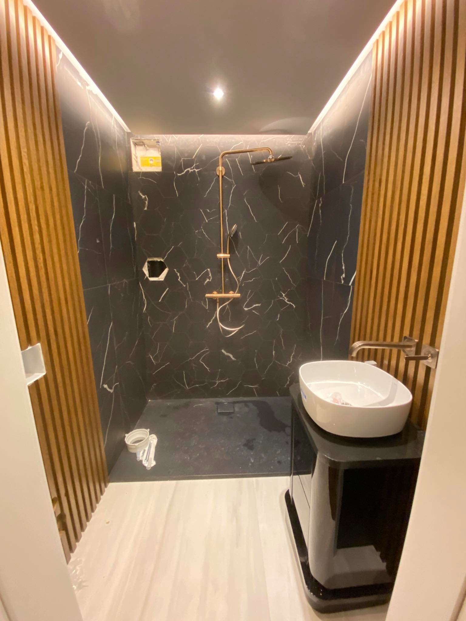 Nowoczesna łazienka w trakcie wykańczania: czarne marmurowe płytki, drewniane lamele, złota armatura prysznicowa, biała umywalka na czarnej szafce, widoczne niedokończone prace przy instalacji...