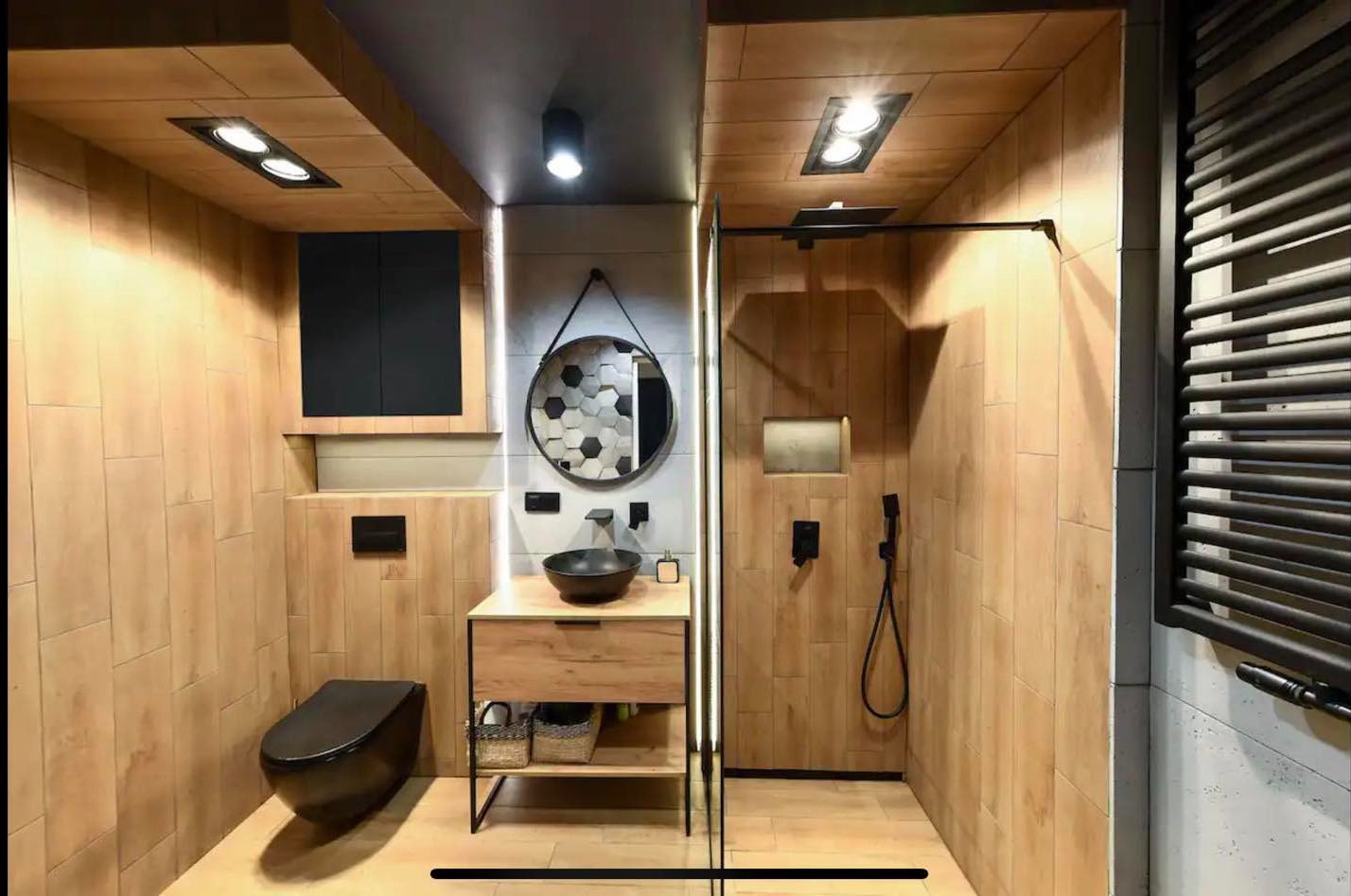 Nowoczesna łazienka z drewnopodobnymi płytkami, czarną armaturą, okrągłym lustrem z heksagonalnym wzorem i kabiną prysznicową typu walk-in.