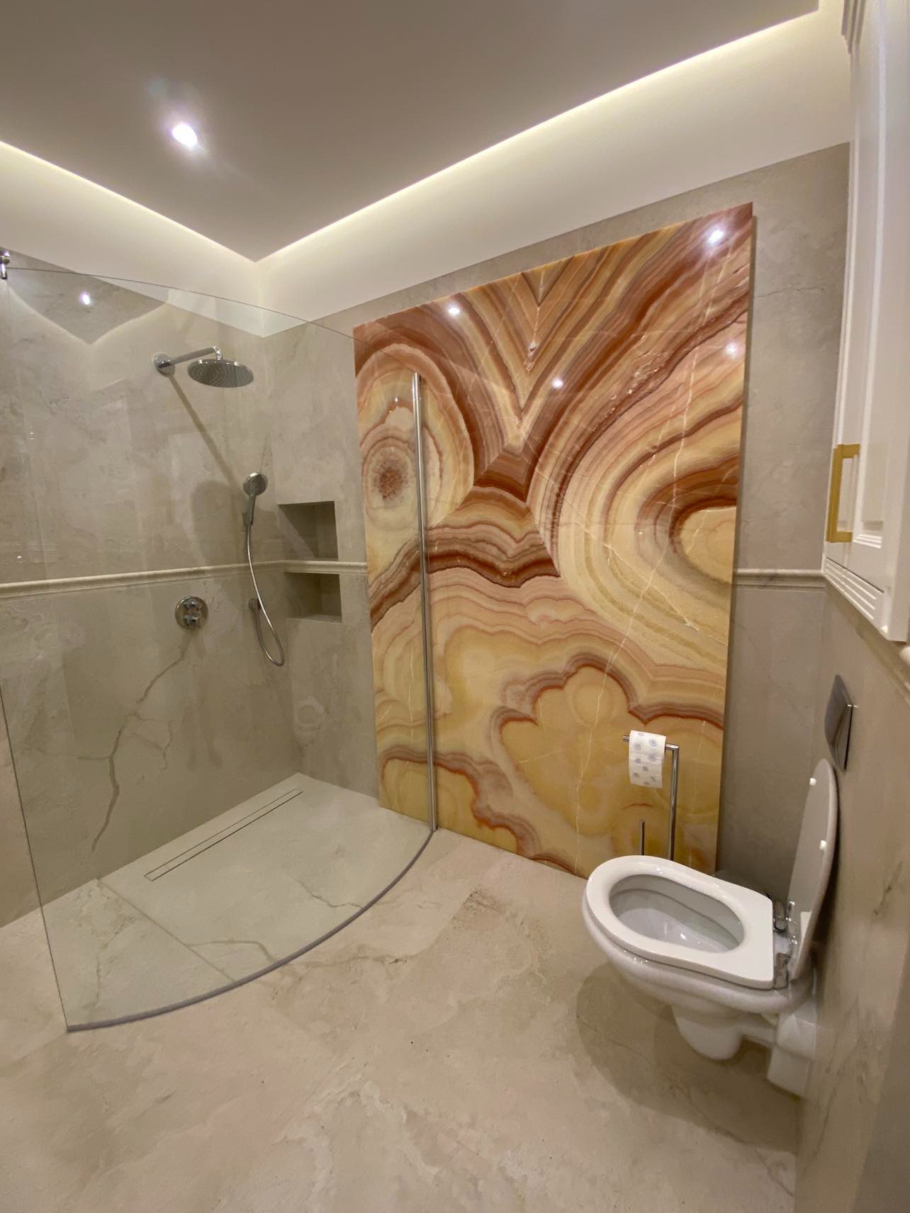 Elegancka łazienka z kabiną prysznicową typu walk-in, wykończona płytami imitującymi marmur oraz dekoracyjnym panelem z onyksu podświetlonym od góry.