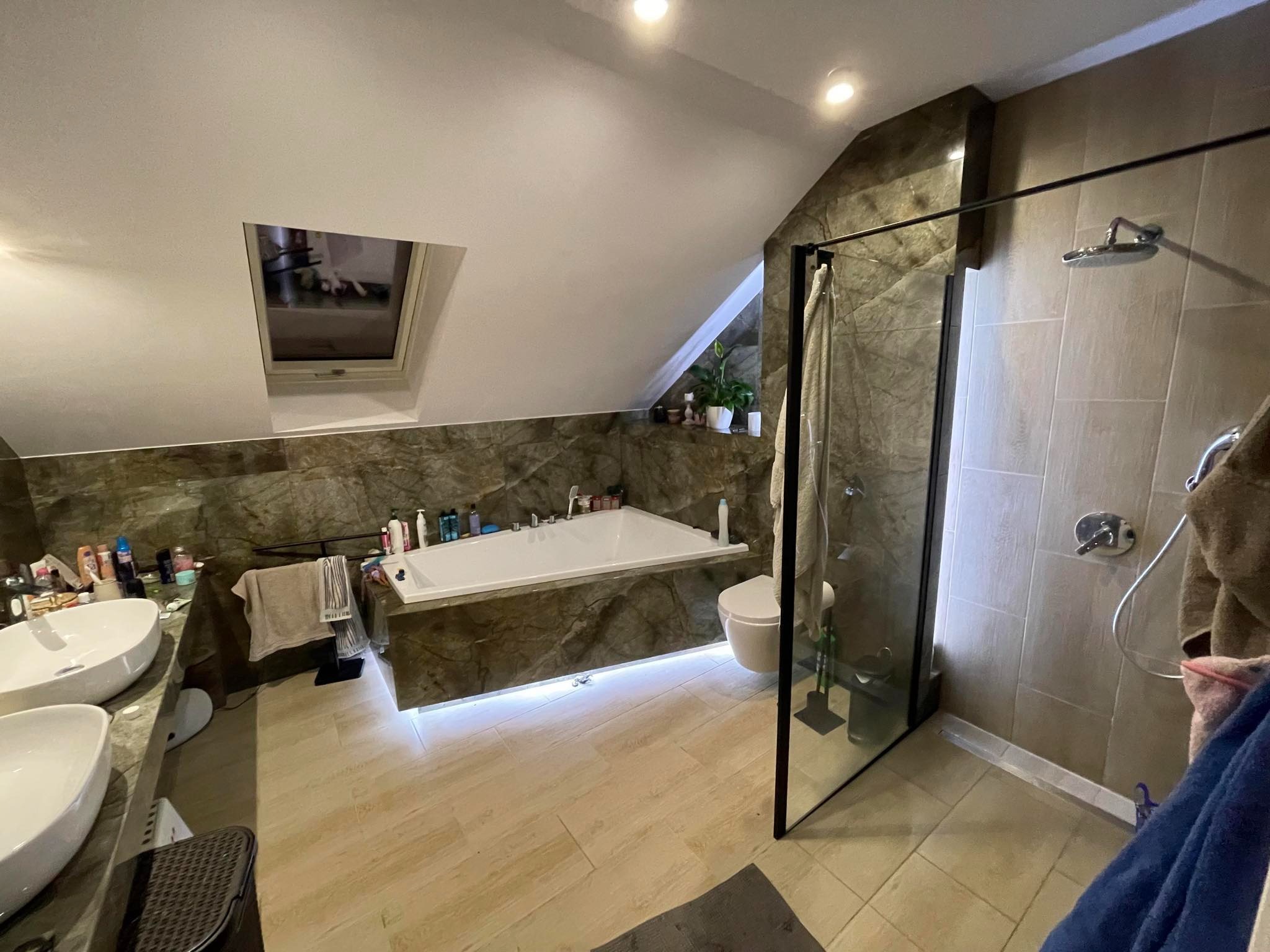 Przestronna łazienka na poddaszu z oknem dachowym, marmurowymi płytkami, wanną z hydromasażem i kabiną prysznicową typu walk-in z czarną ramą.