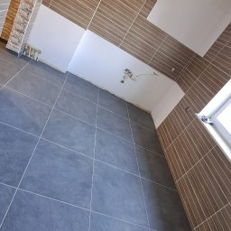 Patryk cz - Wykończone wnętrze łazienki z szarymi płytkami podłogowymi i brązowymi płytkami ściennymi ułożonymi w pionowe pasy, widoczne niedokończone fragmenty sufitu i instalacji elektrycznej.