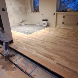 Patryk cz - Montaż drewnianego parkietu w trakcie remontu mieszkania, widoczne narzędzia i materiały budowlane, częściowo wykończona podłoga, przygotowana do dalszych prac wykończeniowych.
