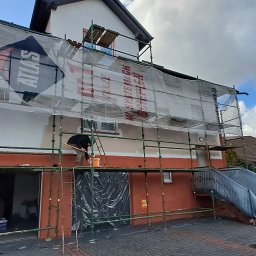 Patryk cz - Dom jednorodzinny w trakcie ocieplania, z widocznym rusztowaniem, siatką ochronną z logo Atlas oraz pracownikiem wykonującym prace na wysokości.