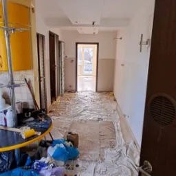 Przygotowanie do remontu: podłoga w korytarzu zabezpieczona folią ochronną z nadrukiem, widoczne narzędzia i materiały malarskie, żółte szafki kuchenne w tle, ściany częściowo pomalowane na biało.