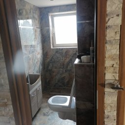 Patryk cz - Wykończona łazienka z brązowo-szarymi płytkami imitującymi marmur na ścianach i podłodze, widoczna umywalka z szafką, toaleta i okno. Dekoracyjne wykończenie ościeżnicy z kamienia elewacyjnego.