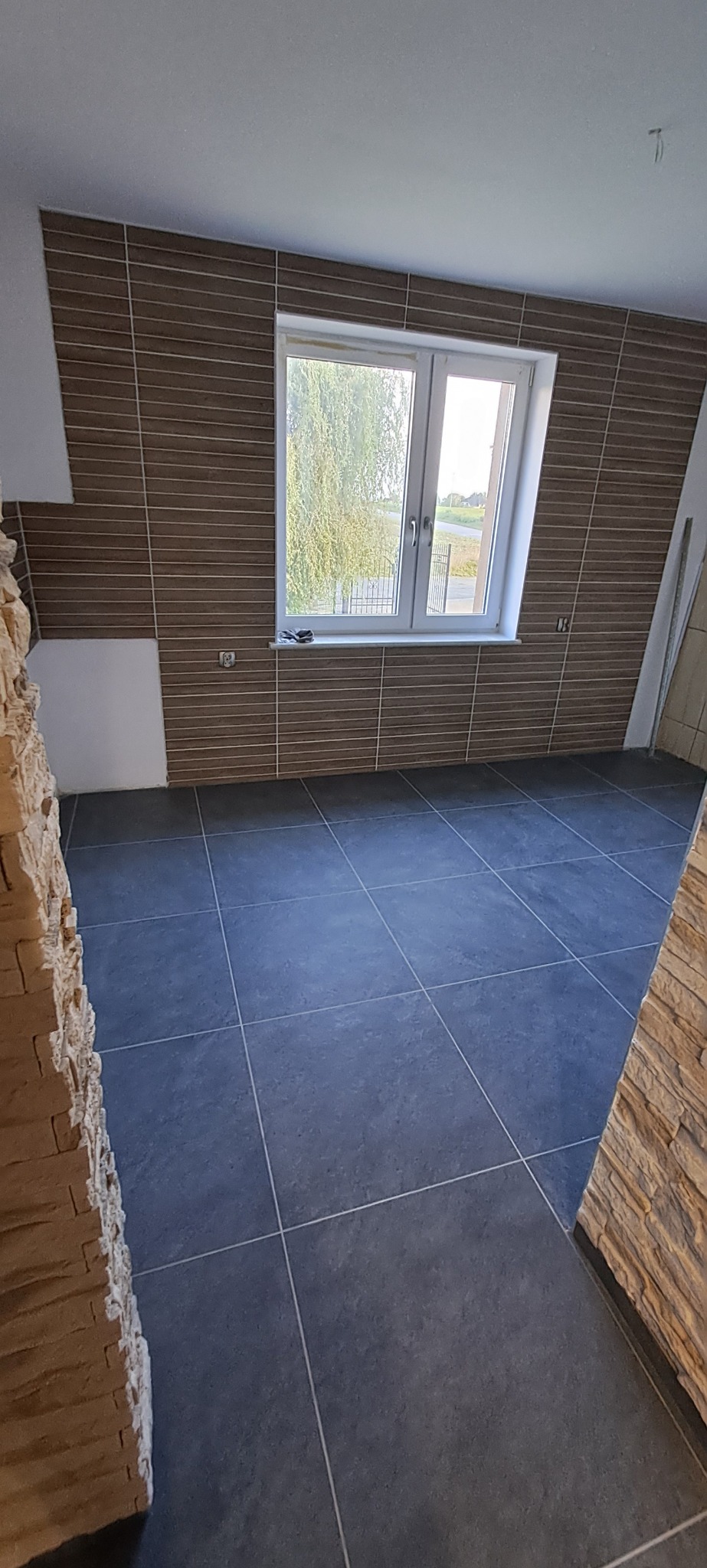 Ukończona łazienka z brązowymi płytkami imitującymi drewno na ścianach, szarą podłogą z płytek i oknem z widokiem na zieleń.