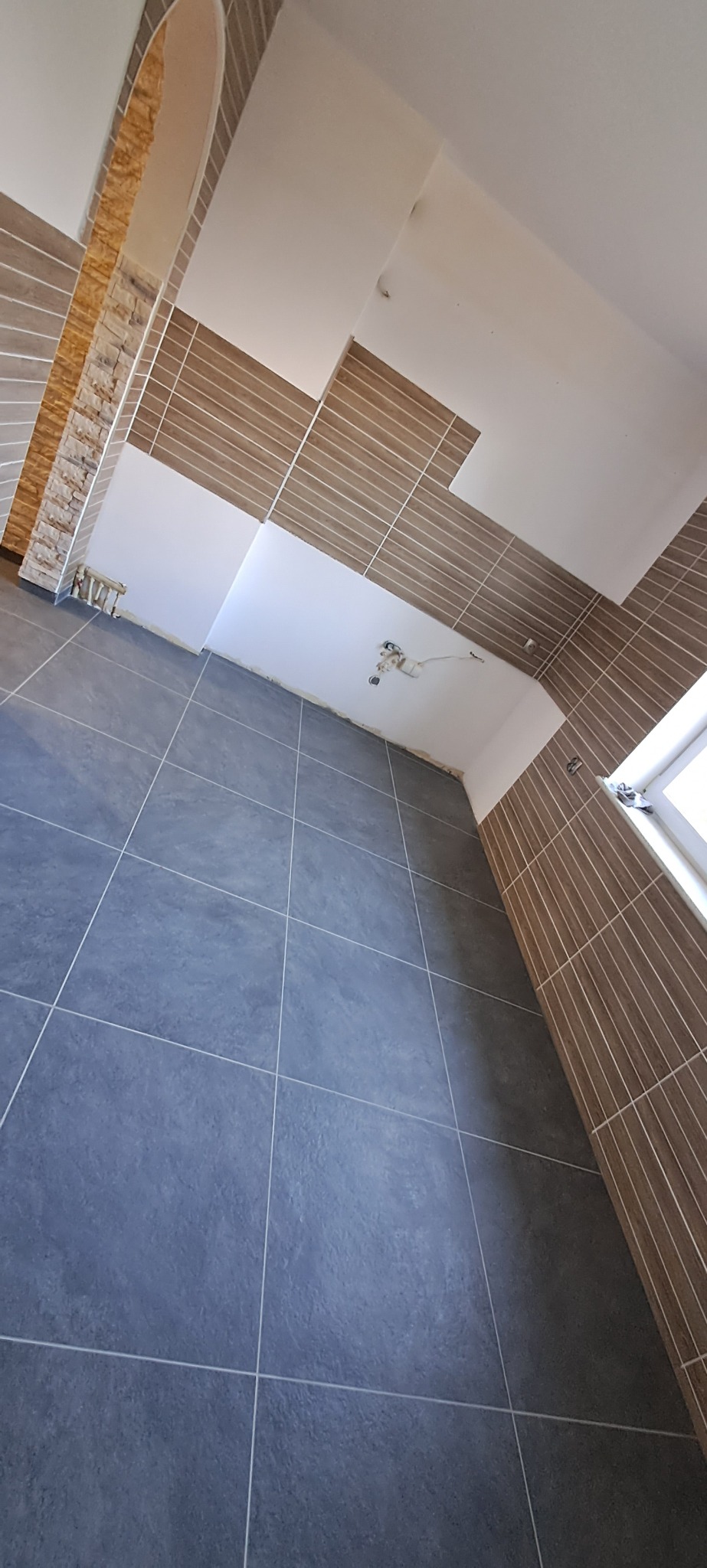 Wykończone wnętrze łazienki z szarymi płytkami podłogowymi i brązowymi płytkami ściennymi ułożonymi w pionowe pasy, widoczne niedokończone fragmenty sufitu i instalacji elektrycznej.