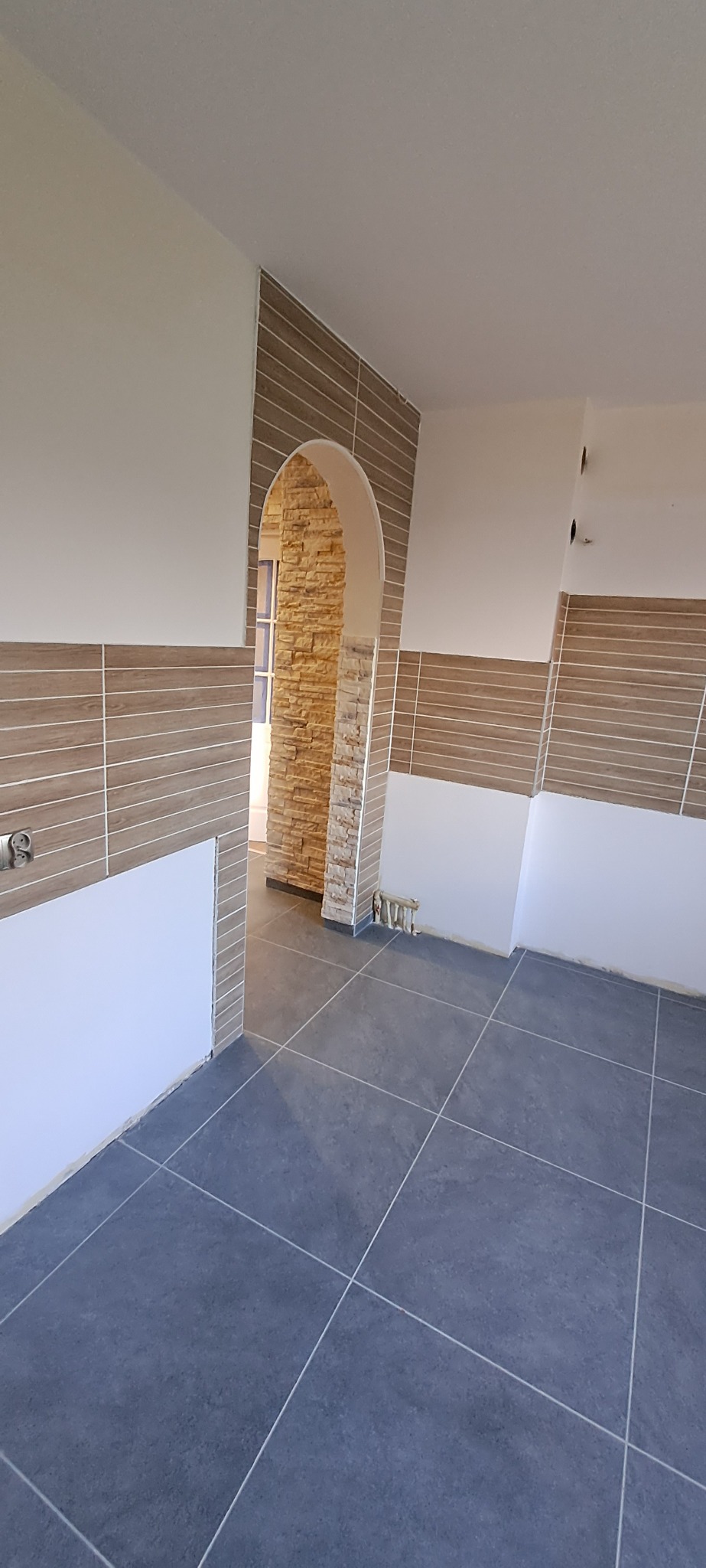 Ukończona łazienka z szarymi płytkami podłogowymi i brązowymi płytkami ściennymi w stylu poziomych paneli, łukowe przejście wykończone kamieniem dekoracyjnym.