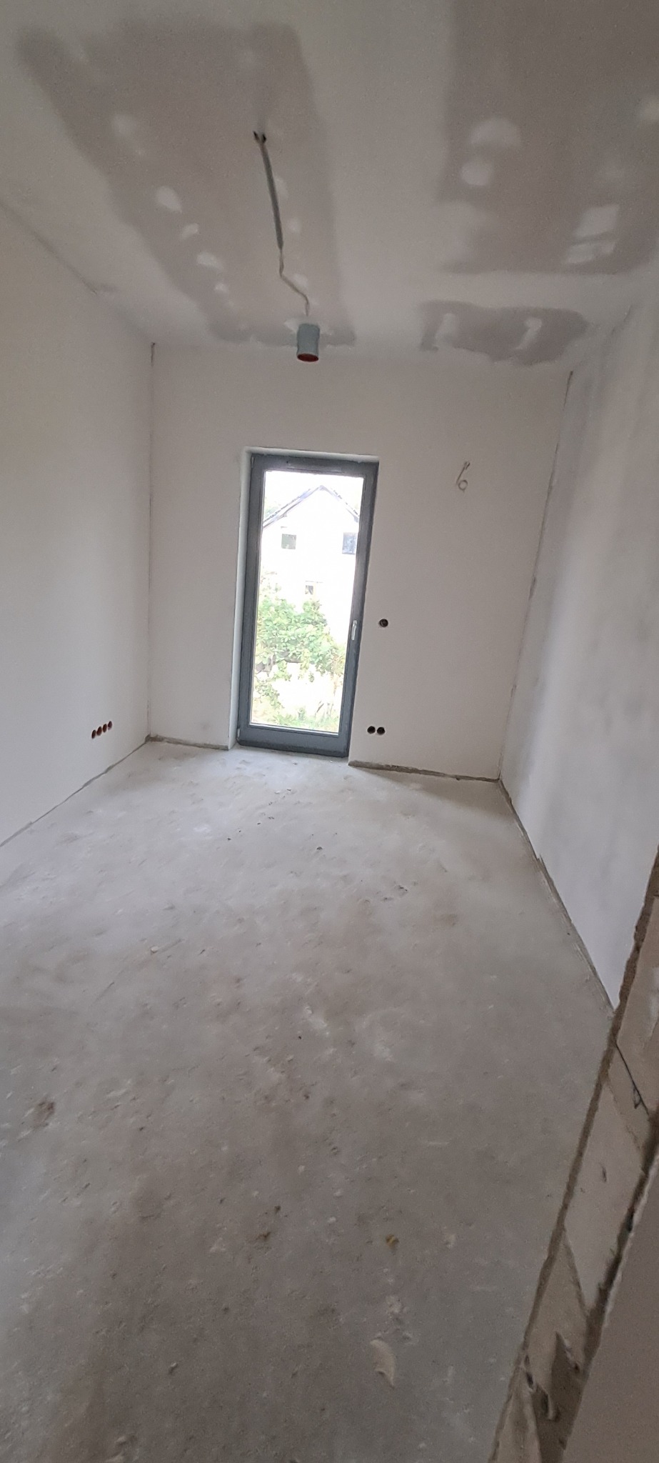 Pokój w trakcie remontu: ściany przygotowane do malowania, widoczne ślady po szpachlowaniu na suficie, surowa podłoga, wystające kable elektryczne, okno z widokiem na zieleń.