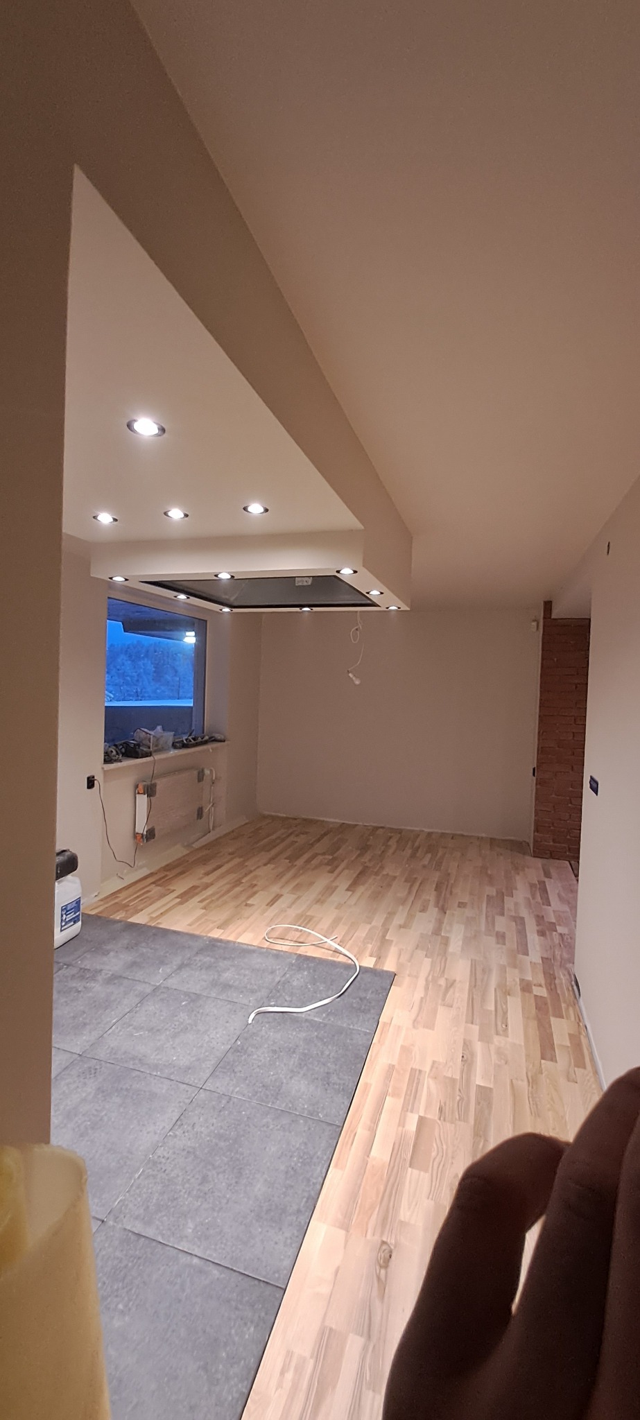 Jasne panele parkietowe ułożone w nowym pomieszczeniu, częściowo przykryte szarymi płytkami, z widoczną instalacją elektryczną i oświetleniem sufitowym.