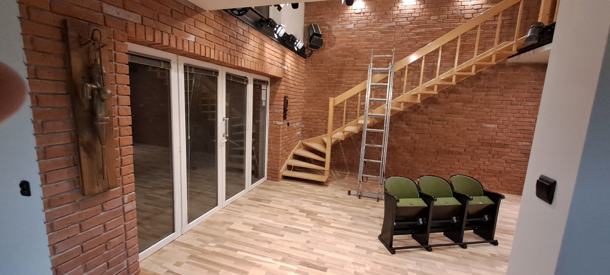 Loftowe wnętrze z ceglaną ścianą, drewnianymi schodami, parkietem i trzema kinowymi fotelami z zielonymi siedziskami; widoczna metalowa drabina i oświetlenie teatralne na suficie.