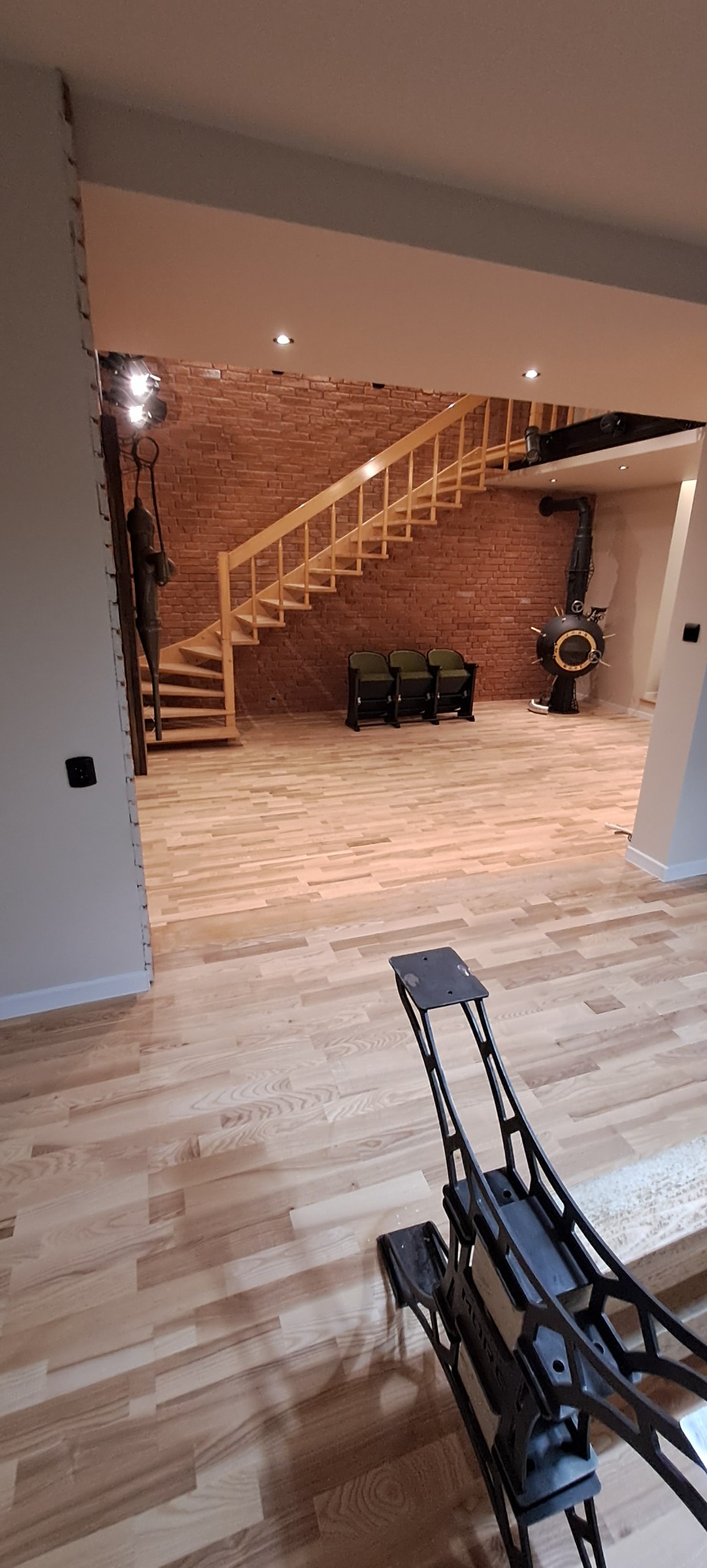 Jasny parkiet ułożony w przestronnym salonie z ceglaną ścianą, drewnianymi schodami i ozdobnym kominkiem; w pierwszym planie narzędzie do układania parkietu.