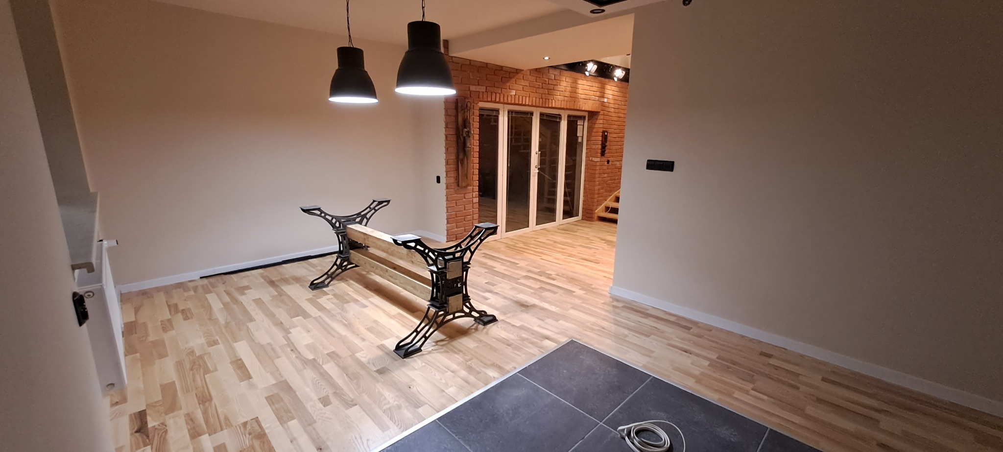 Jasny parkiet ułożony w przestronnym wnętrzu z ceglaną ścianą w tle, widoczne industrialne nogi stołu i wiszące lampy.