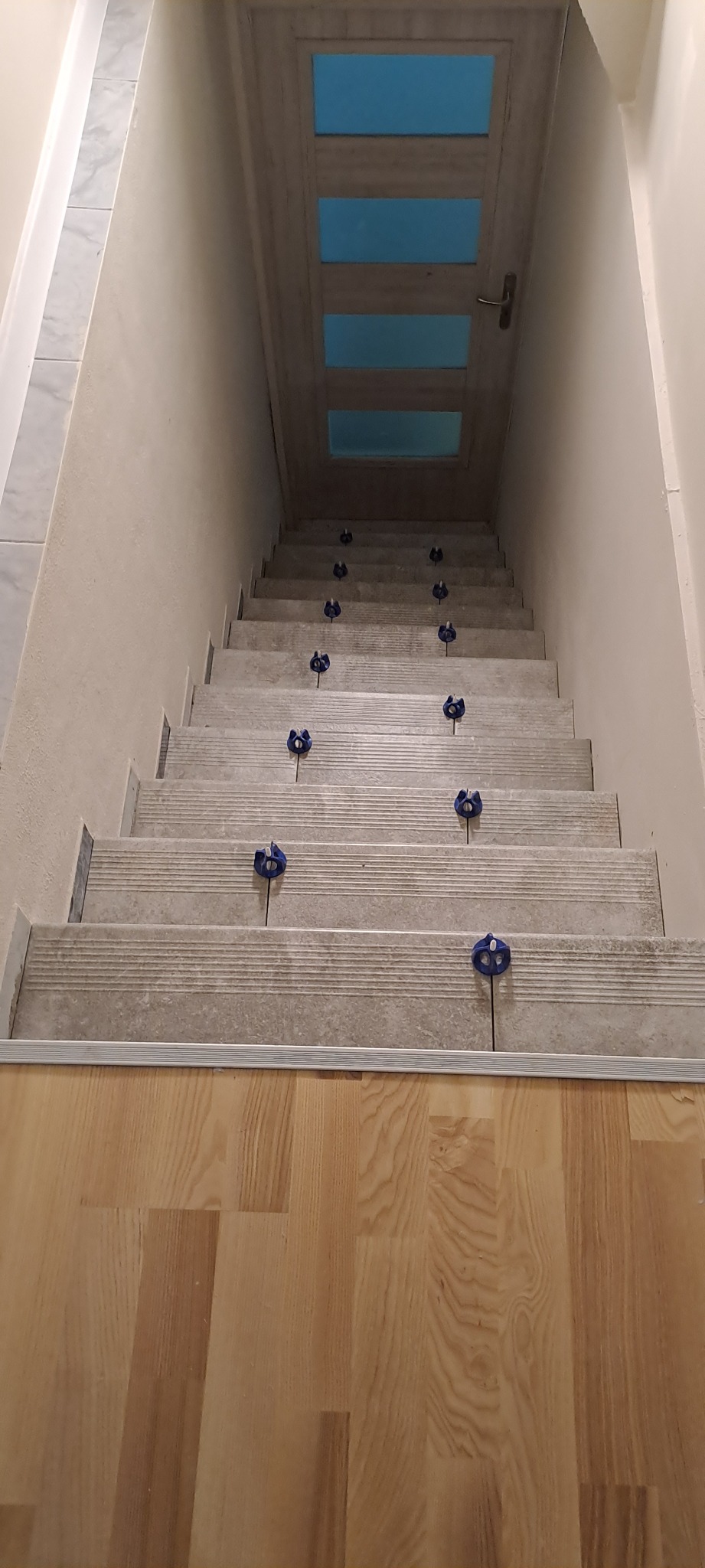 Schody wyłożone płytkami imitującymi kamień, z niebieskimi krzyżykami dystansowymi, prowadzące do drzwi z szybami w odcieniach błękitu, widok z góry na dół.