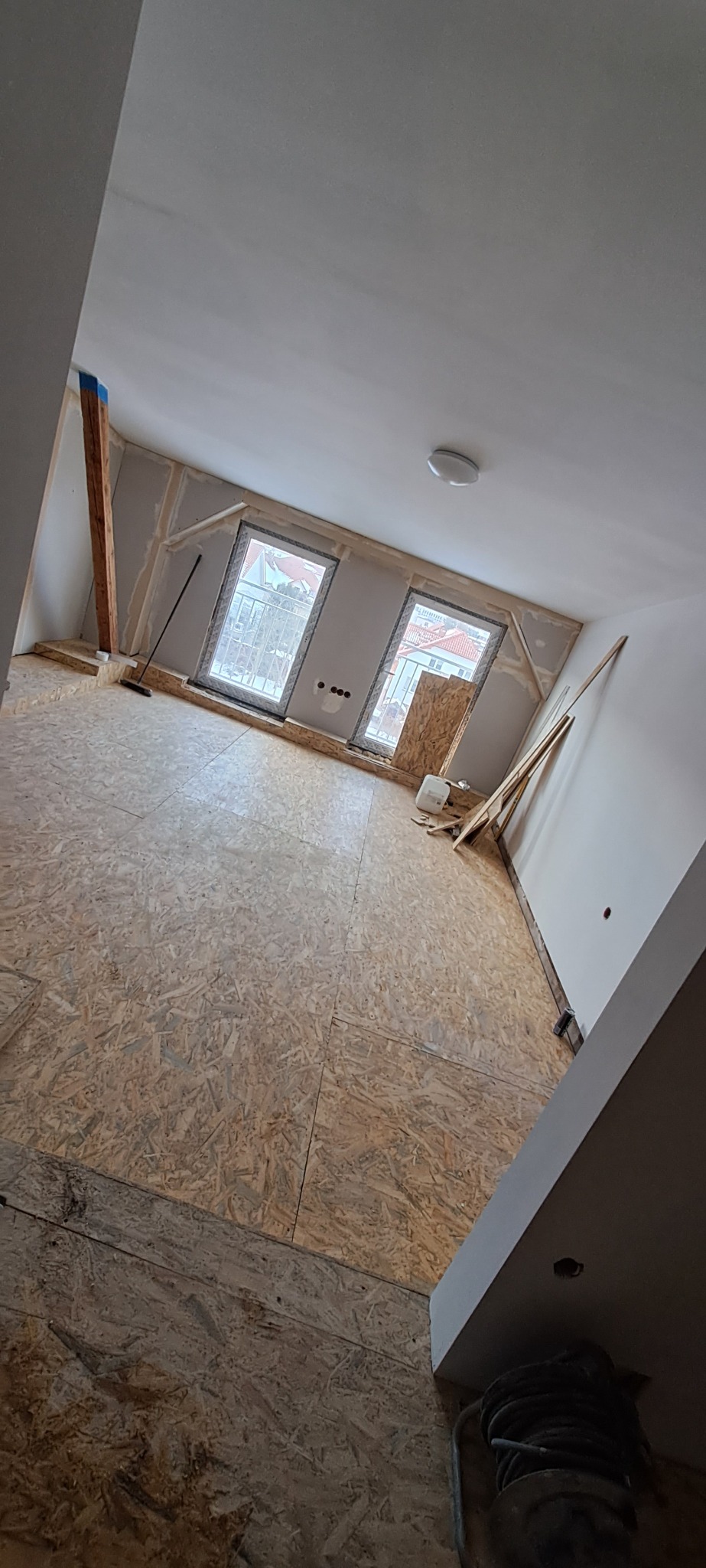 Poddasze w trakcie adaptacji: widoczne płyty OSB na podłodze, ściany przygotowane do szpachlowania, okna dachowe z widokiem na zabudowę mieszkalną, drewniany słup wzmacniający konstrukcję.