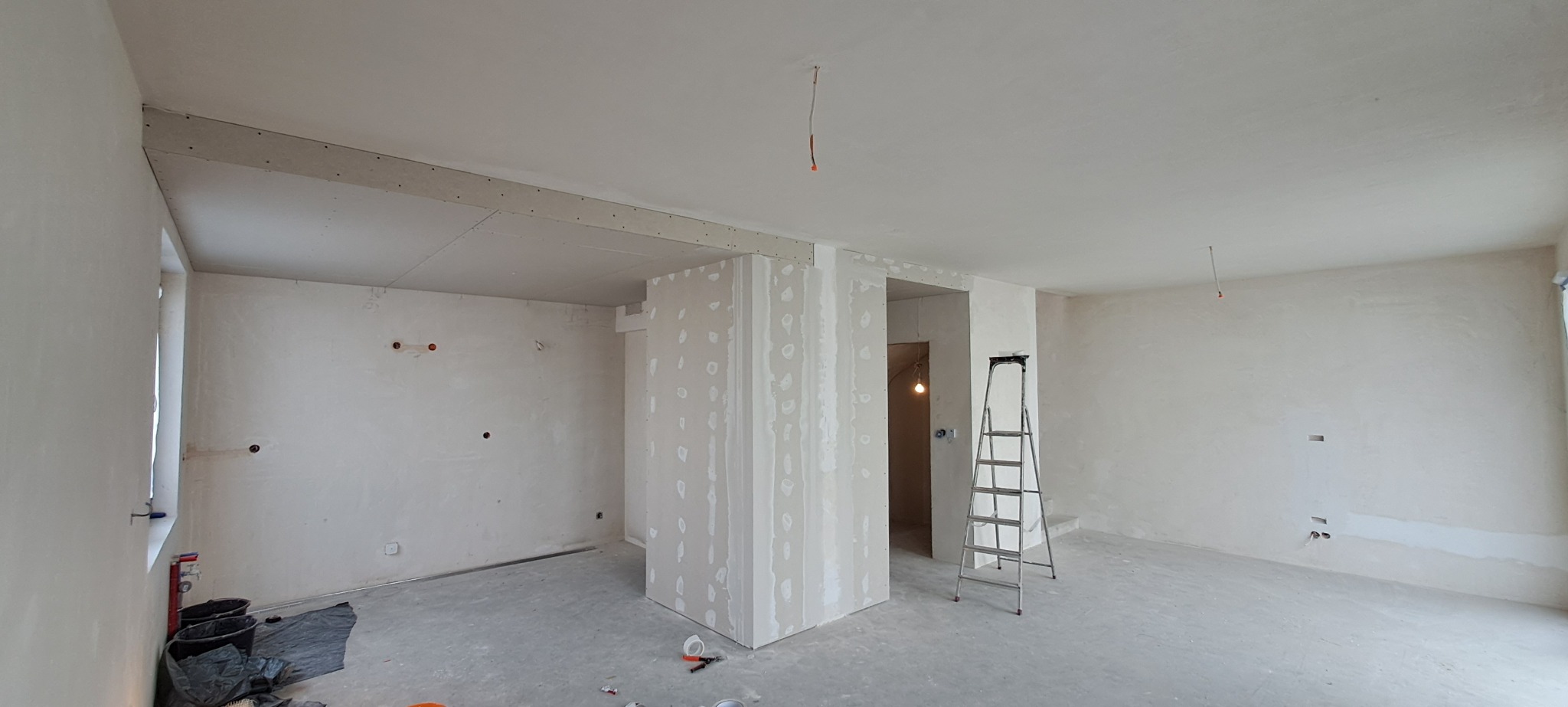Wnętrze w trakcie prac wykończeniowych: ściany i sufit pokryte gładzią szpachlową, widoczne łączenia płyt gipsowo-kartonowych, drabina aluminiowa w pomieszczeniu, przygotowane otwory pod instalacje...
