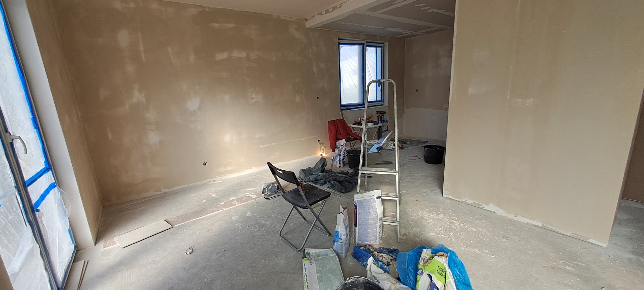 Pokój w trakcie szpachlowania: ściany pokryte warstwą szpachli, okno zabezpieczone folią, drabina aluminiowa, worki ze szpachlą i narzędzia malarskie rozłożone na podłodze.