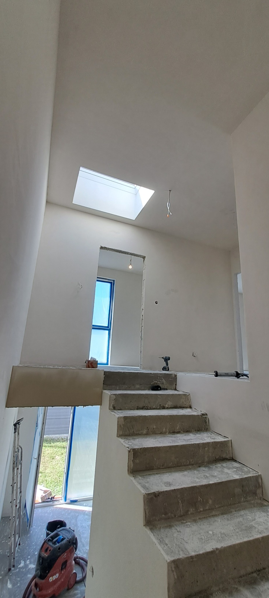 Wnętrze w trakcie remontu: surowe schody betonowe prowadzą na górę, ściany i sufit po szpachlowaniu, widoczne okno dachowe i okno na dole zabezpieczone folią, narzędzia budowlane na schodach...