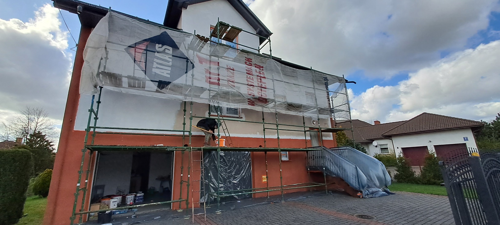 Dom jednorodzinny w trakcie ocieplania, z widocznym rusztowaniem, siatką ochronną z logo Atlas oraz pracownikiem wykonującym prace na wysokości.