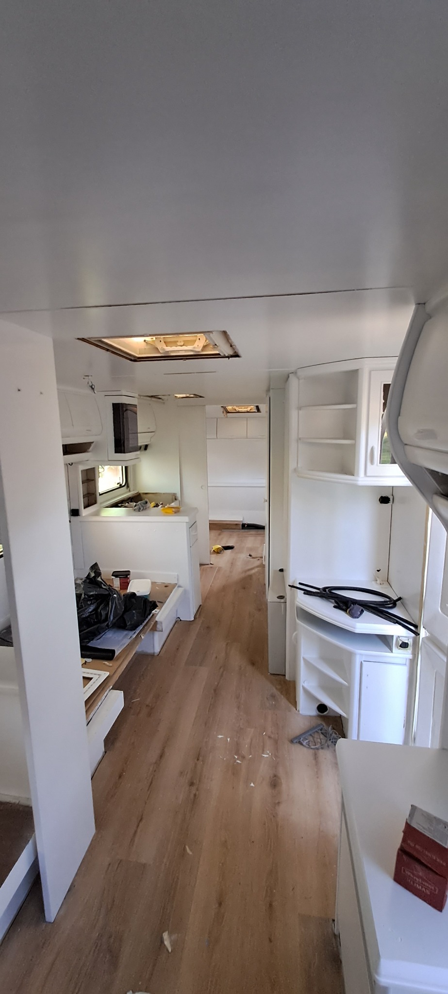Wnętrze remontowanego kampera z nową podłogą imitującą drewno i świeżo pomalowanymi na biało ścianami i meblami, widoczne narzędzia i śmieci wskazują na trwający remont.