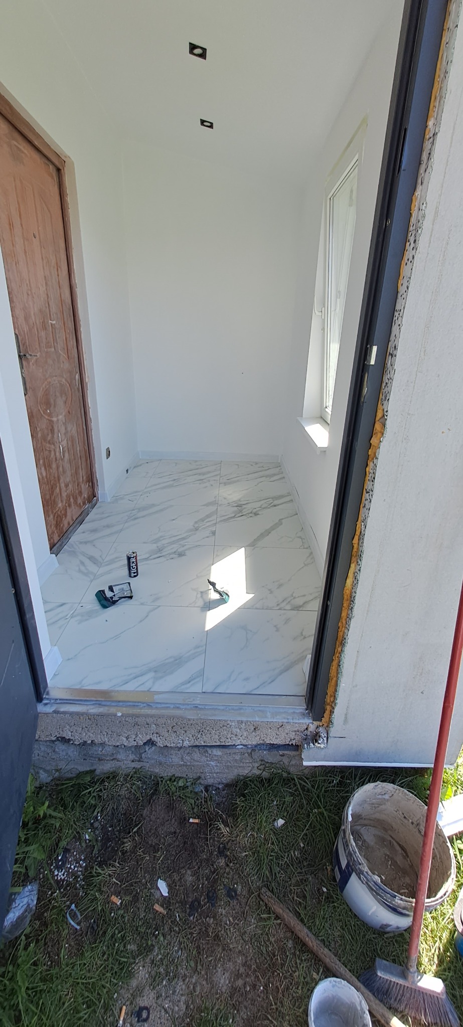 Wnętrze w trakcie remontu: położona marmurowa podłoga z widocznymi narzędziami, puszką po napoju energetycznym i niedokończonym wykończeniem ościeżnicy, na zewnątrz resztki materiałów budowlanych...