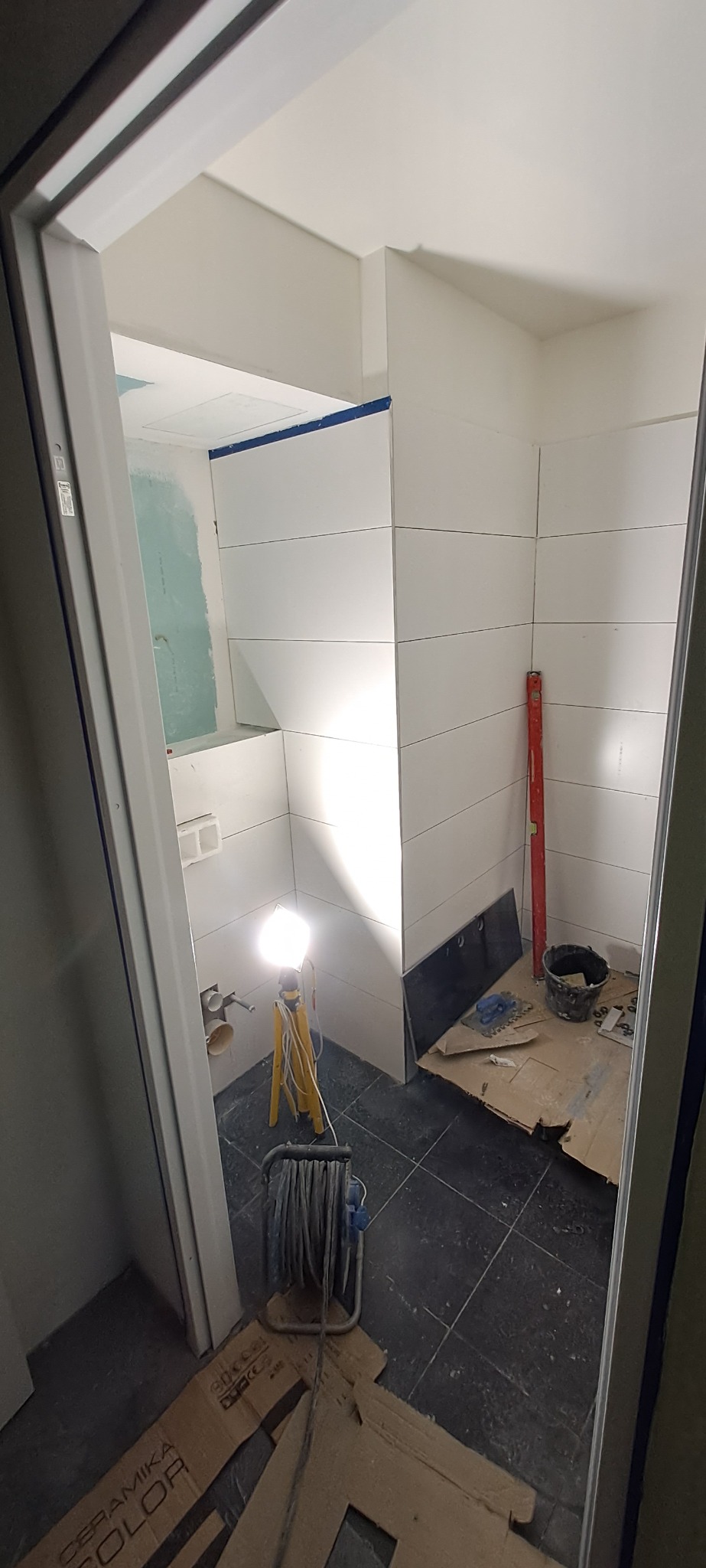 Łazienka w trakcie remontu: białe płytki ułożone poziomo na ścianach, podłoga z ciemnych płytek, widoczne narzędzia, poziomica, wiadro i lampa budowlana na trójnogu, zabezpieczenia na ścianach...