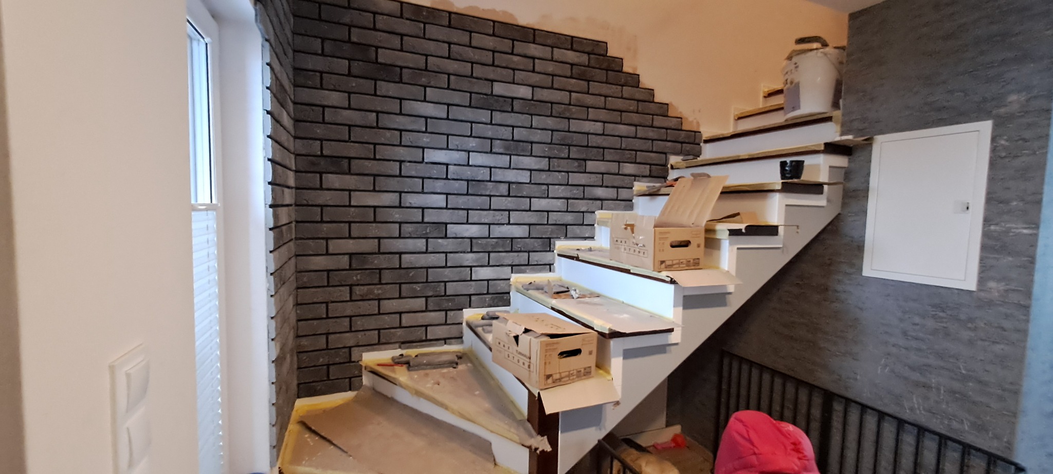 Montaż szarej cegły dekoracyjnej na ścianie przy schodach, widoczne narzędzia i kartony na stopniach, etap prac wykończeniowych.